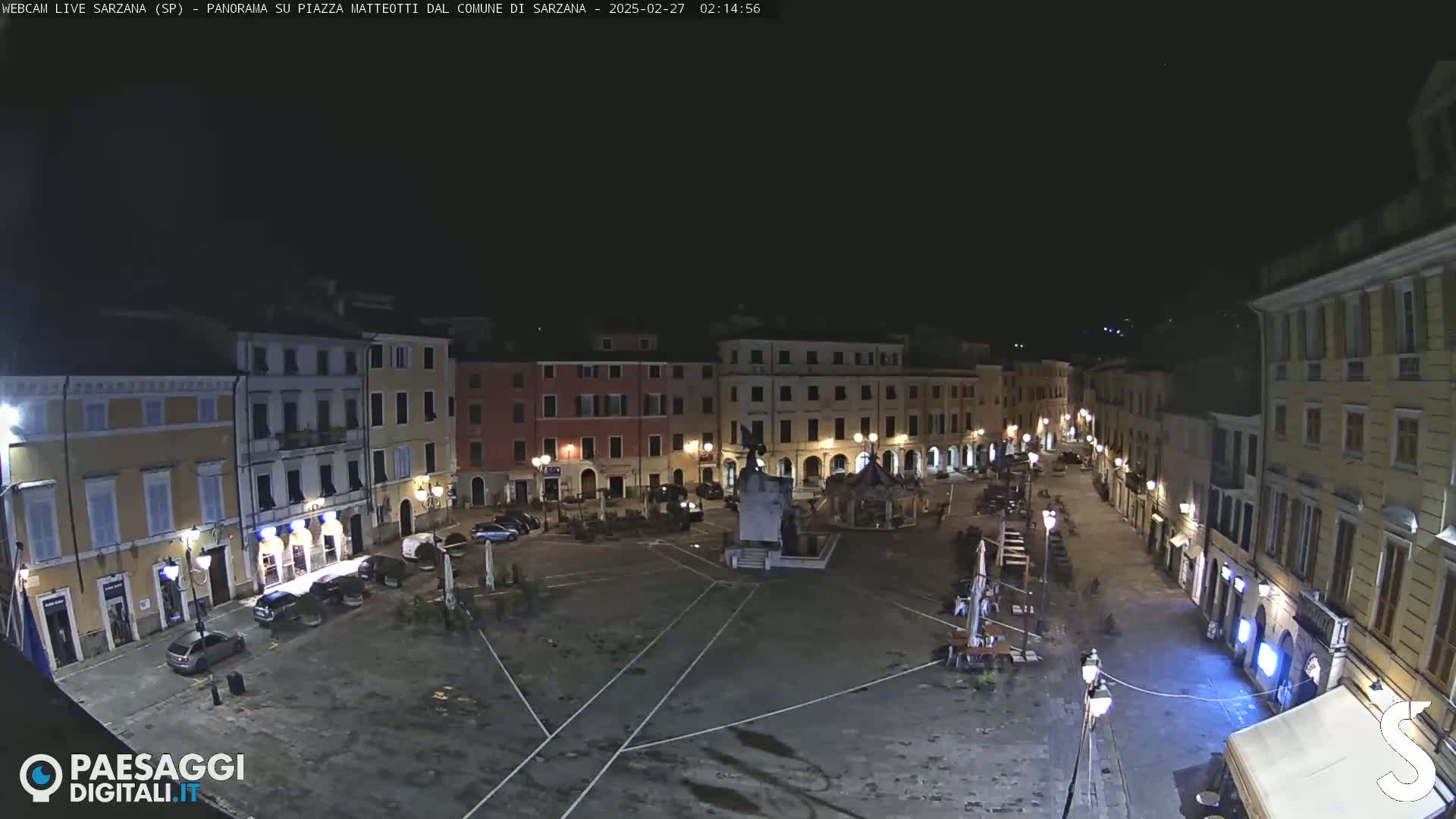 A nighttime, high-angle view of a mostly empty town square surrounded by buildings, with a monument in the center.