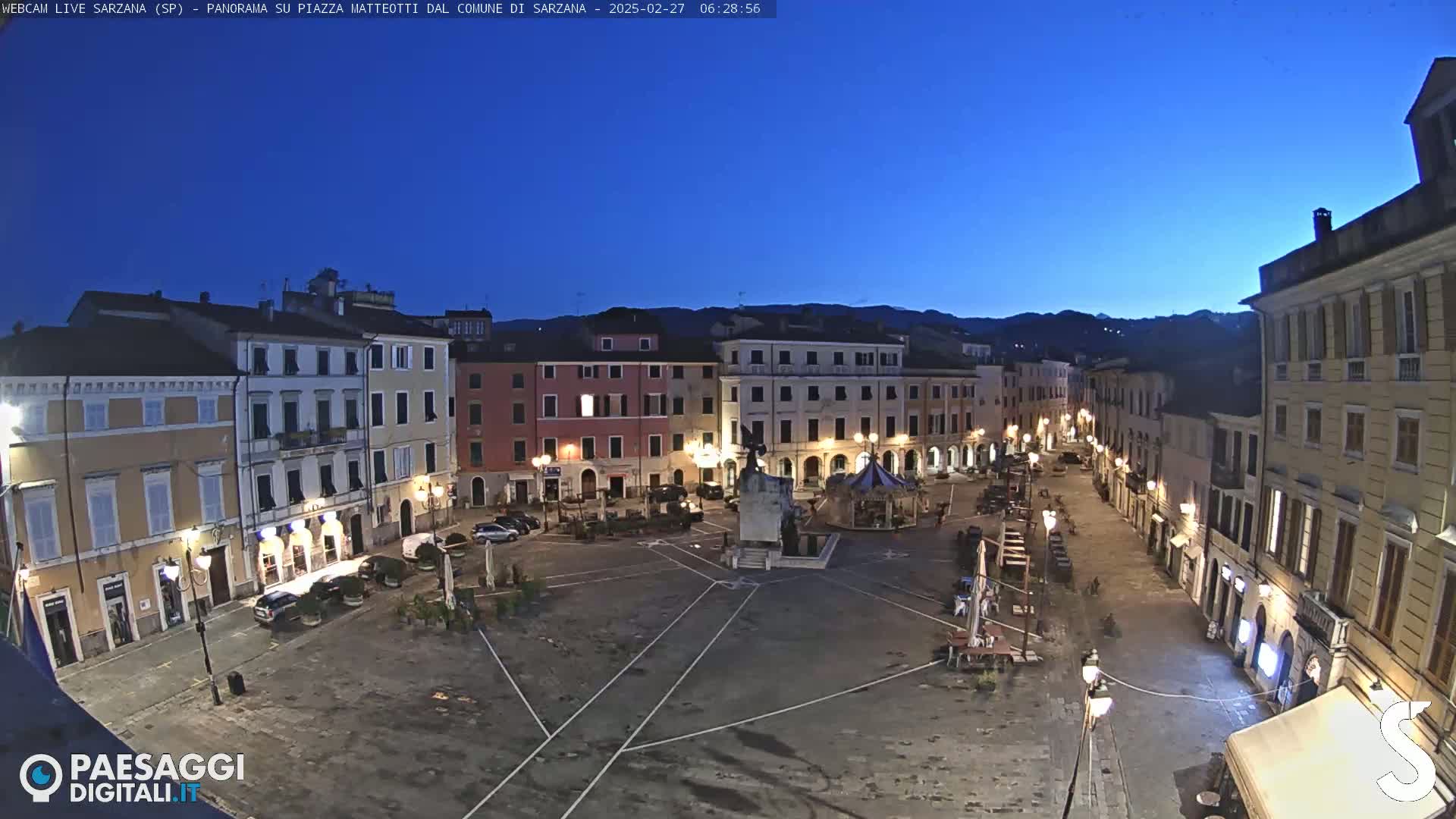 A mostly empty town square at twilight, surrounded by multi-story buildings, under a clear, dark blue sky.