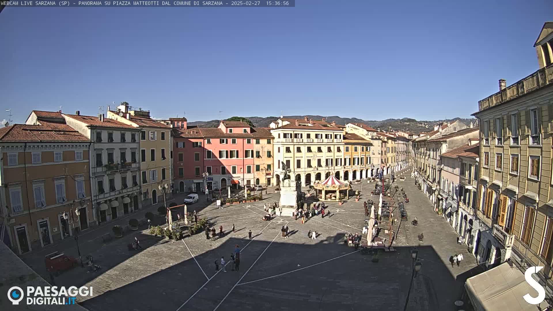 A large town square with several multi-story buildings surrounding it under a clear blue sky.