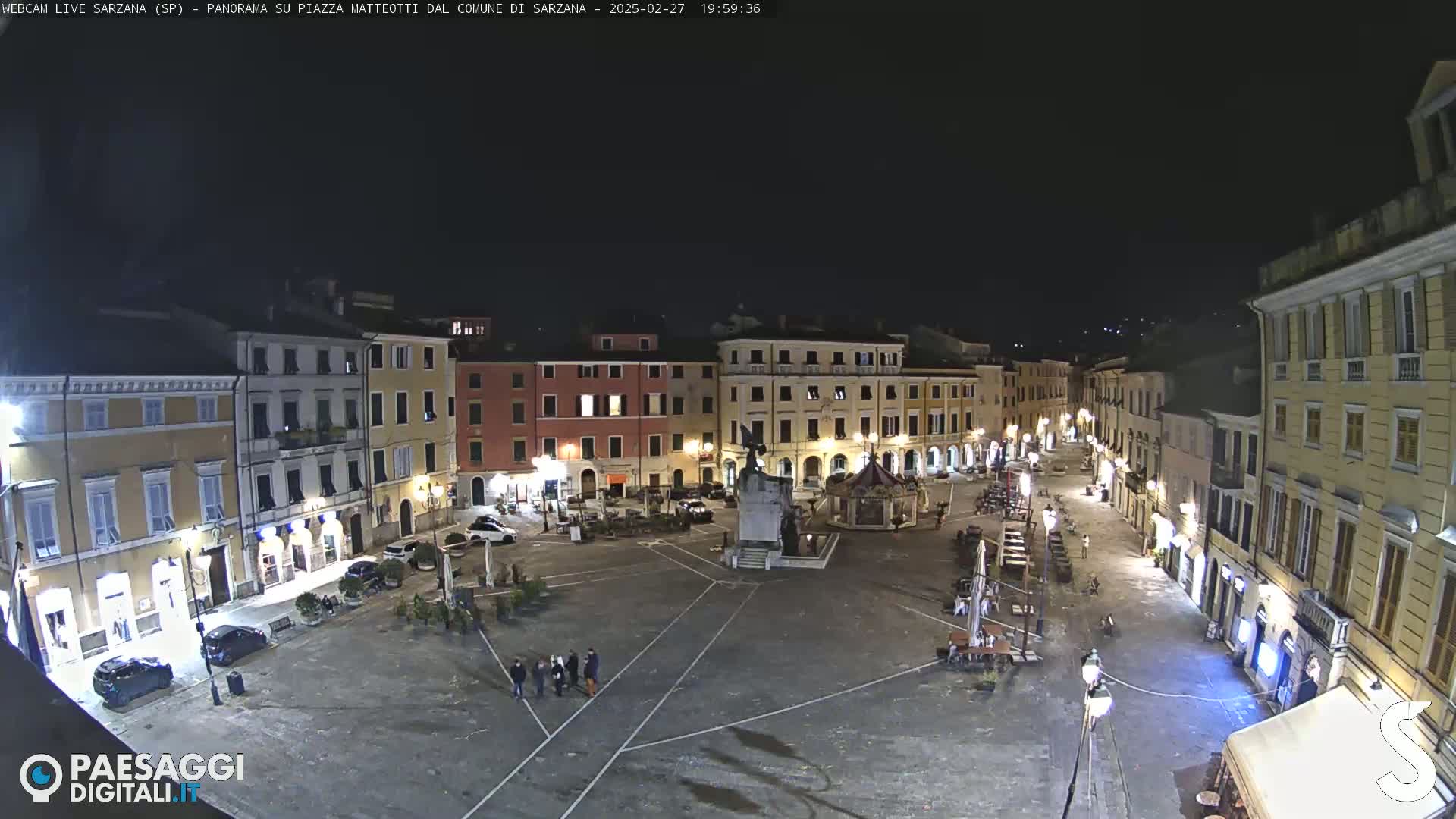 A nighttime, aerial view of a town square with several multi-story buildings surrounding it, sparsely populated with a few people and vehicles.
