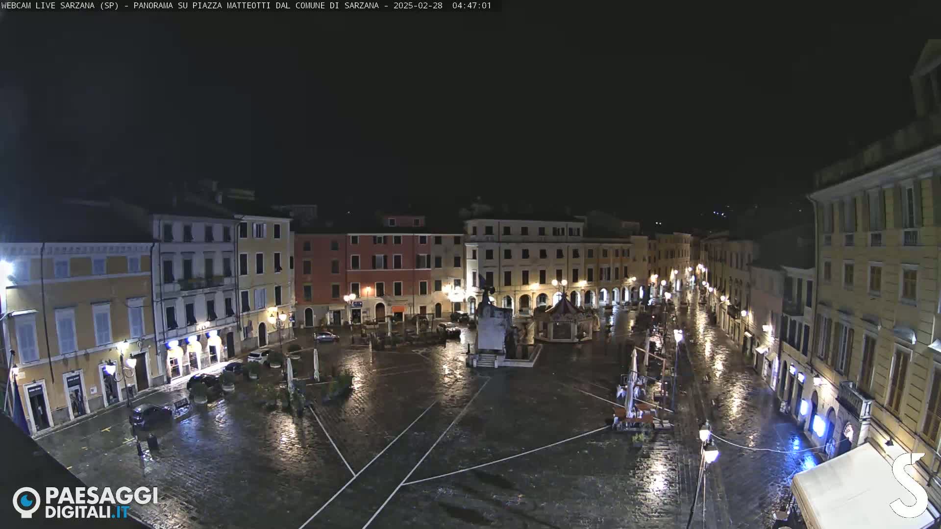 A rain-slicked, nighttime town square is surrounded by illuminated buildings.