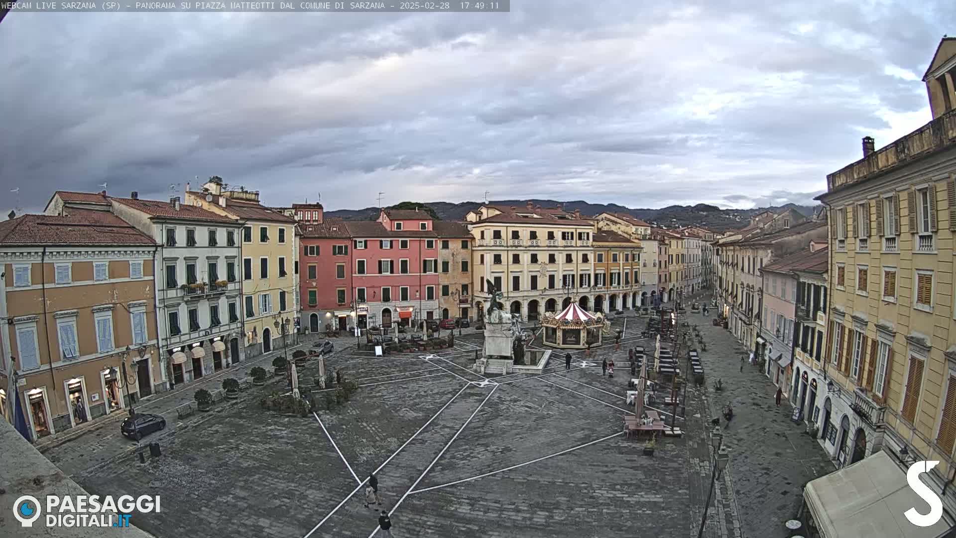 A mostly empty town square, under a cloudy sky, is surrounded by multi-story buildings.