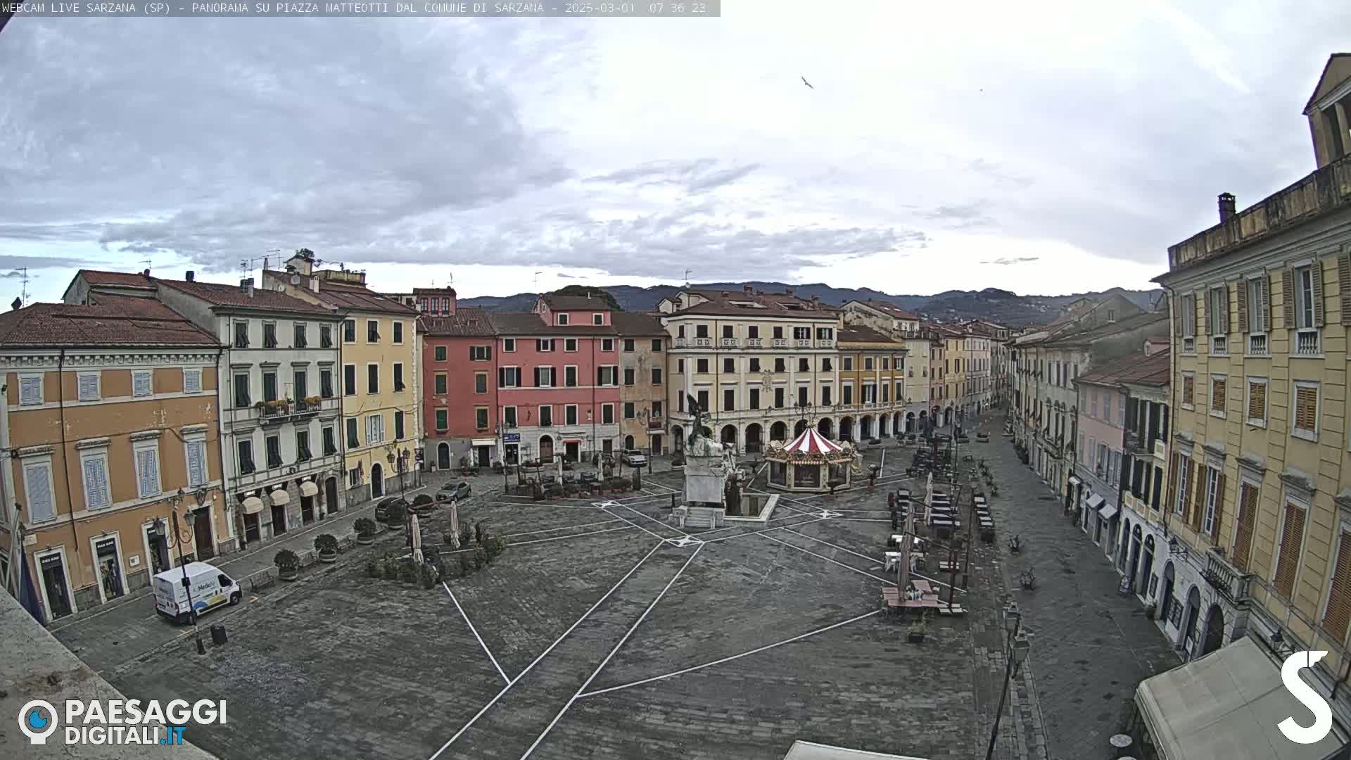 A mostly empty town square with multi-story buildings surrounding it under an overcast sky.