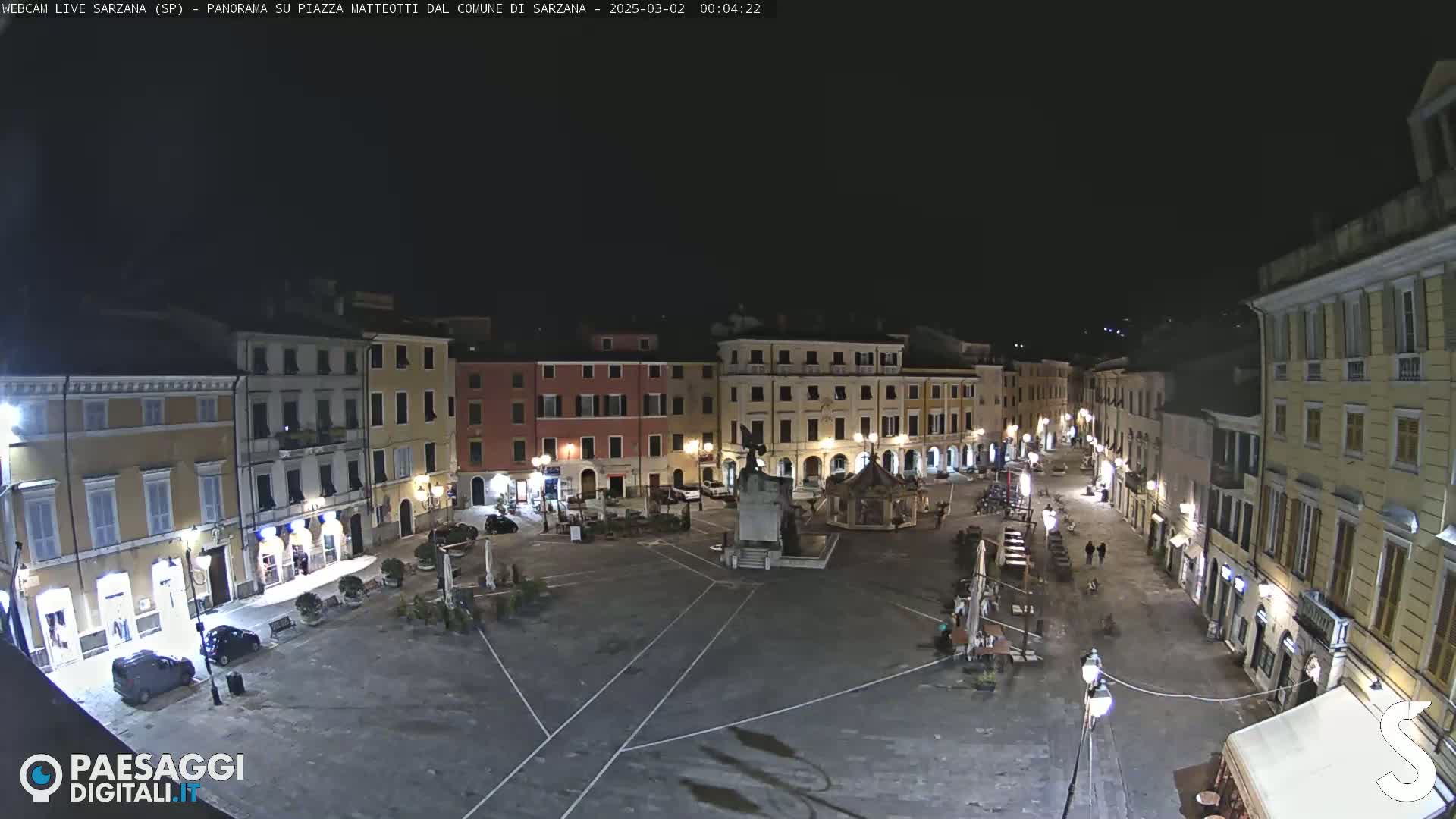 A nighttime, high-angle view of a mostly empty town square surrounded by multi-story buildings with their lights on.