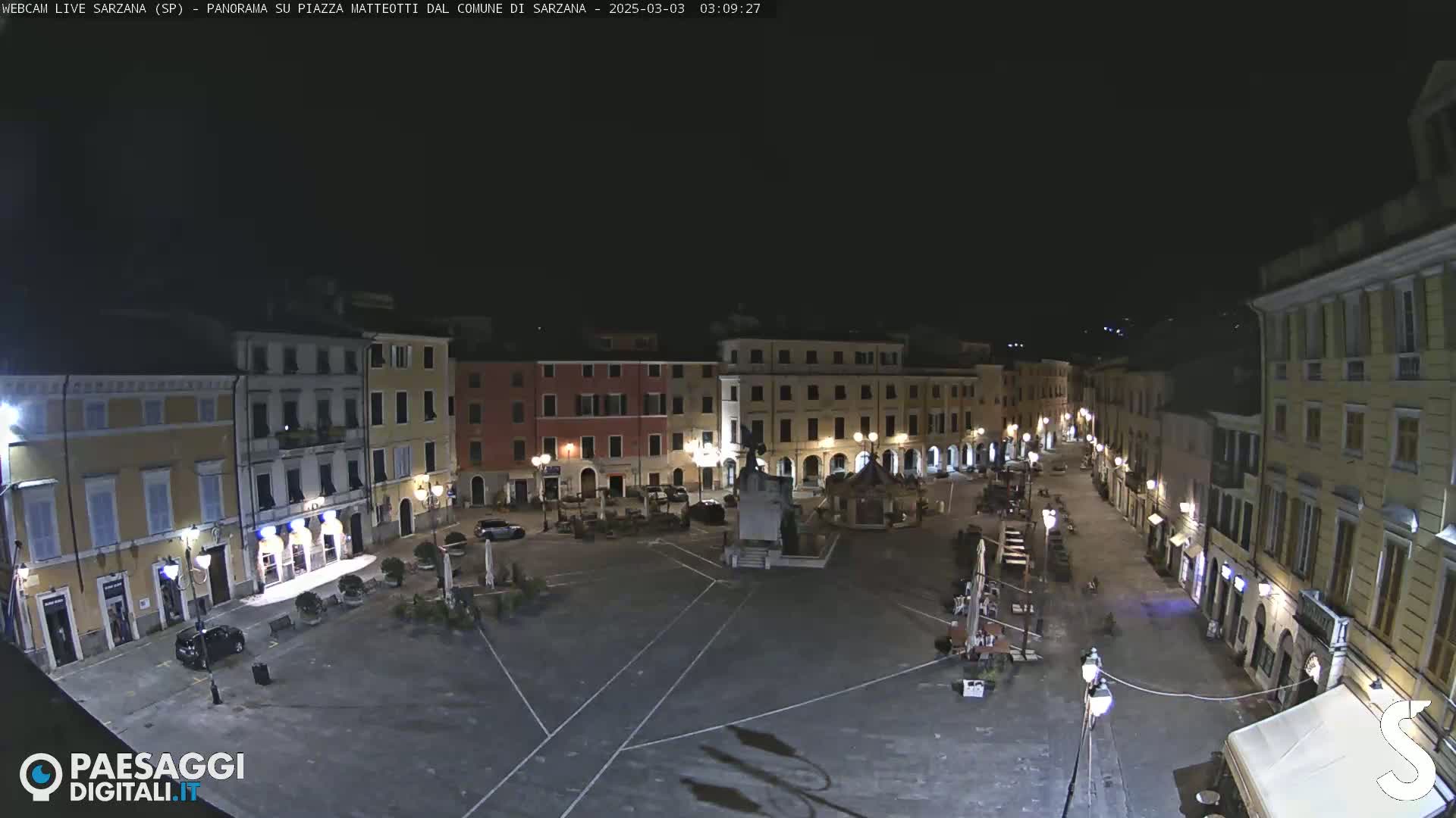 A nighttime, high-angle view of a mostly empty town square surrounded by buildings with illuminated windows and streetlights.