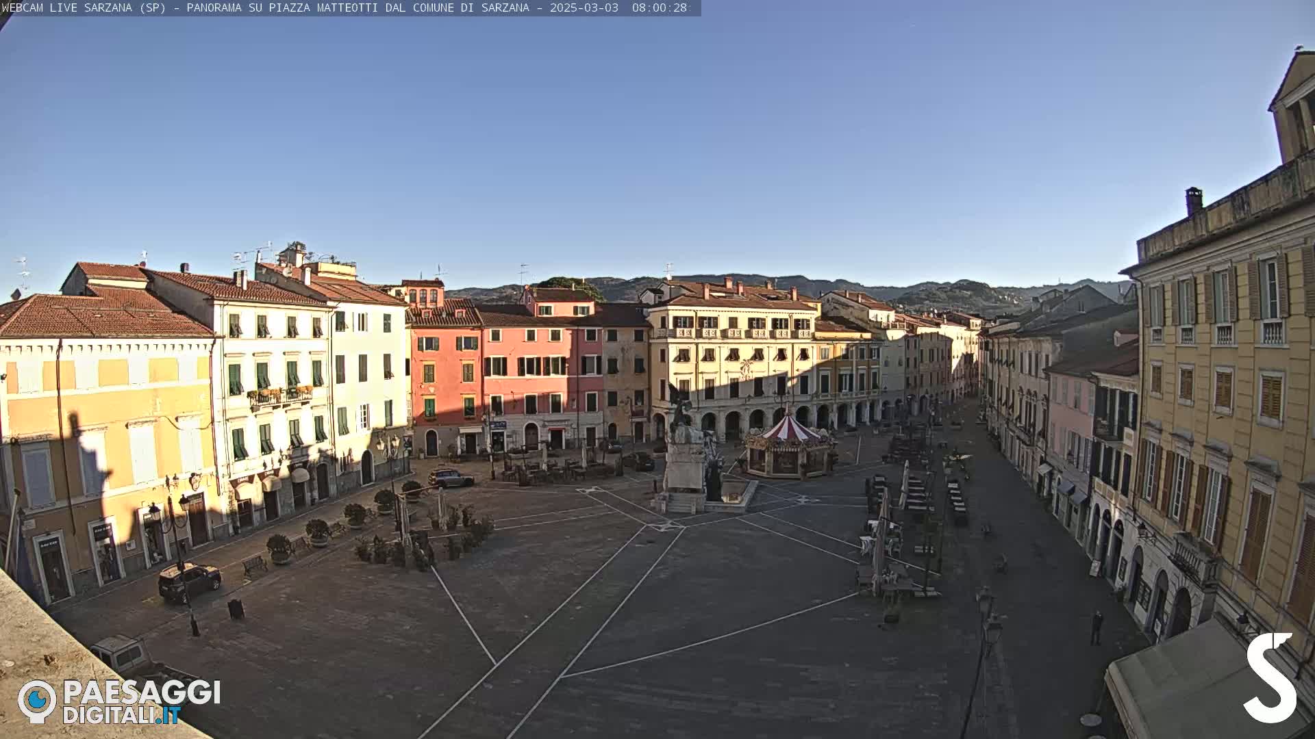 A mostly empty town square, surrounded by multi-story buildings, is bathed in the sunlight of a clear morning.