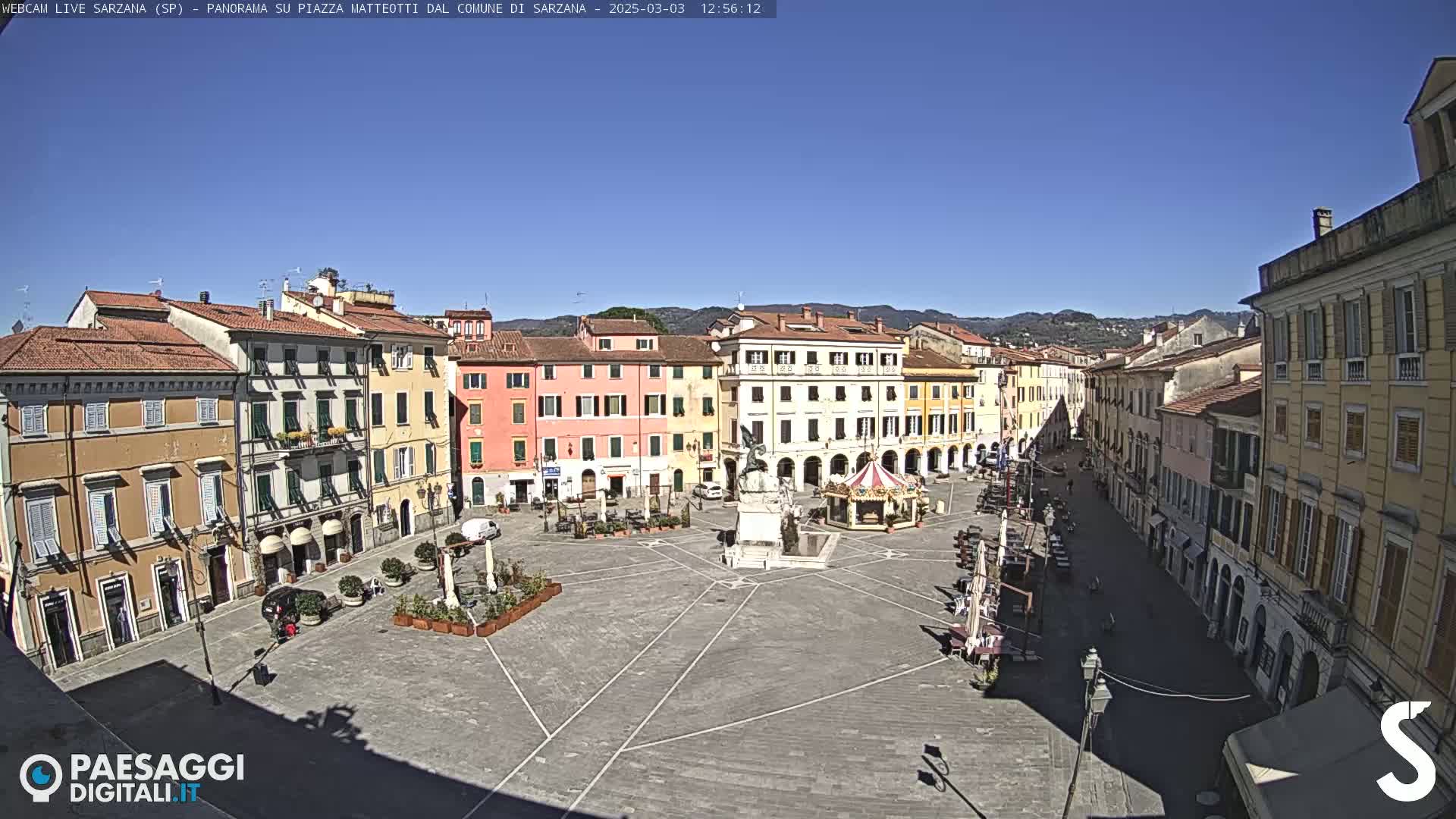 A large, mostly empty town square with pastel-colored buildings surrounding it under a clear blue sky.