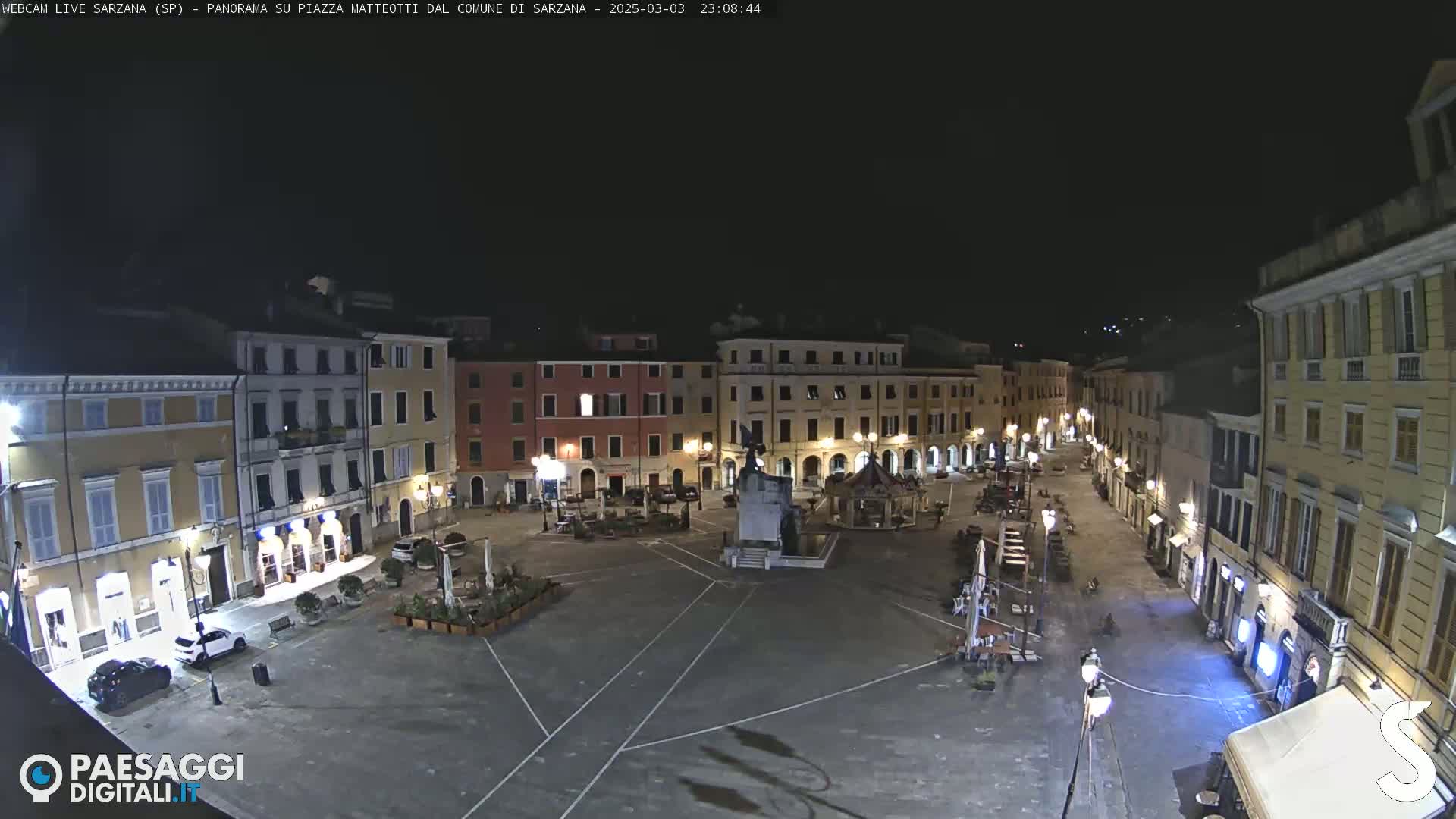 A nighttime, high-angle view of a mostly empty town square, surrounded by buildings with illuminated windows.