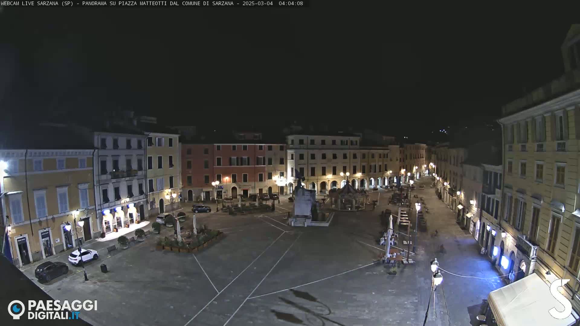 A nighttime view of a town square, illuminated by streetlights, with several buildings lining its perimeter and a few cars parked in the square.