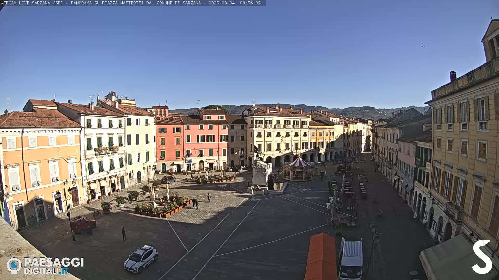 A sunny day reveals a large town square surrounded by pastel-colored buildings, a carousel, and a few parked cars.