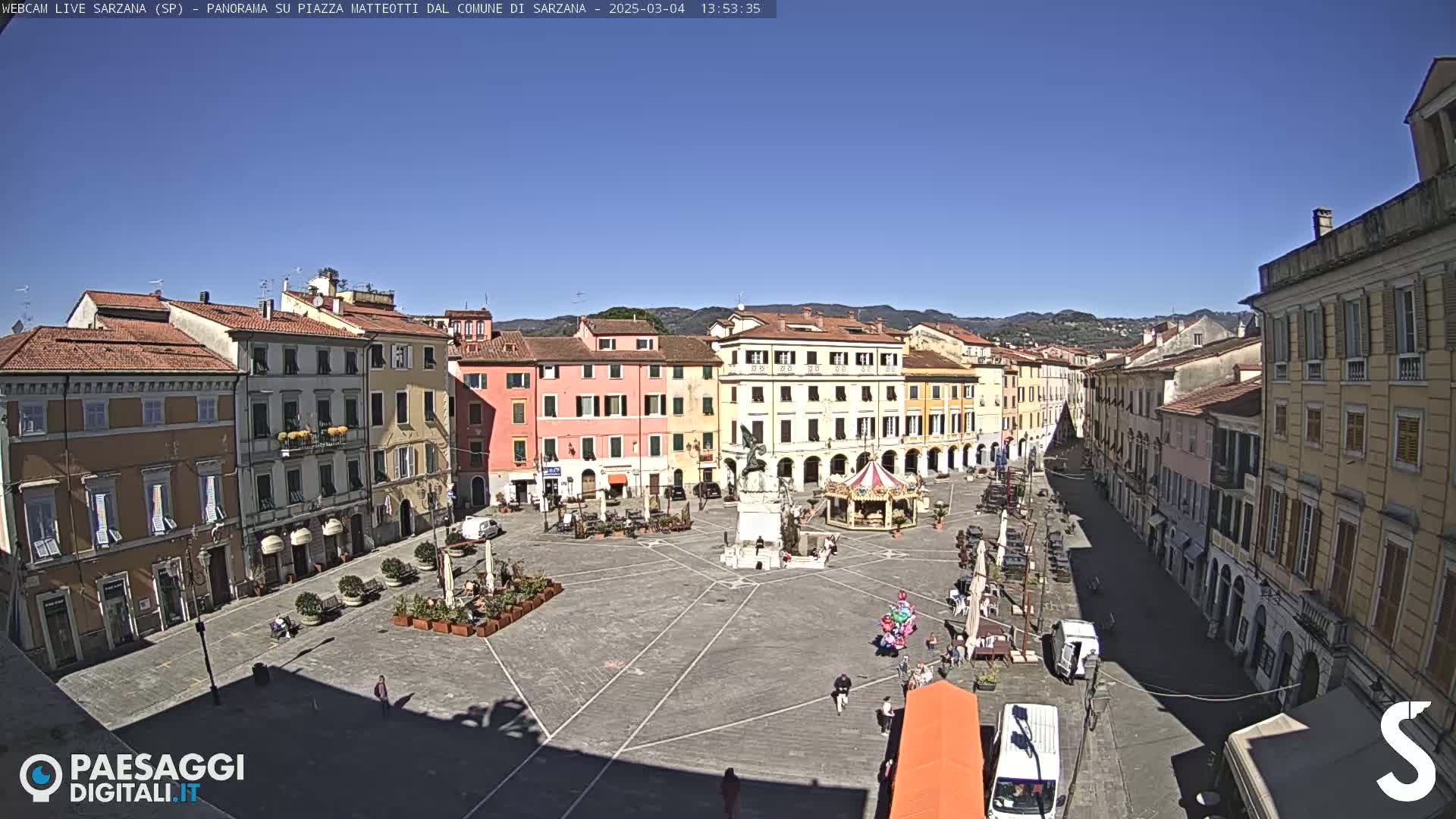 A large, mostly empty town square is surrounded by multi-story buildings under a clear, sunny sky.