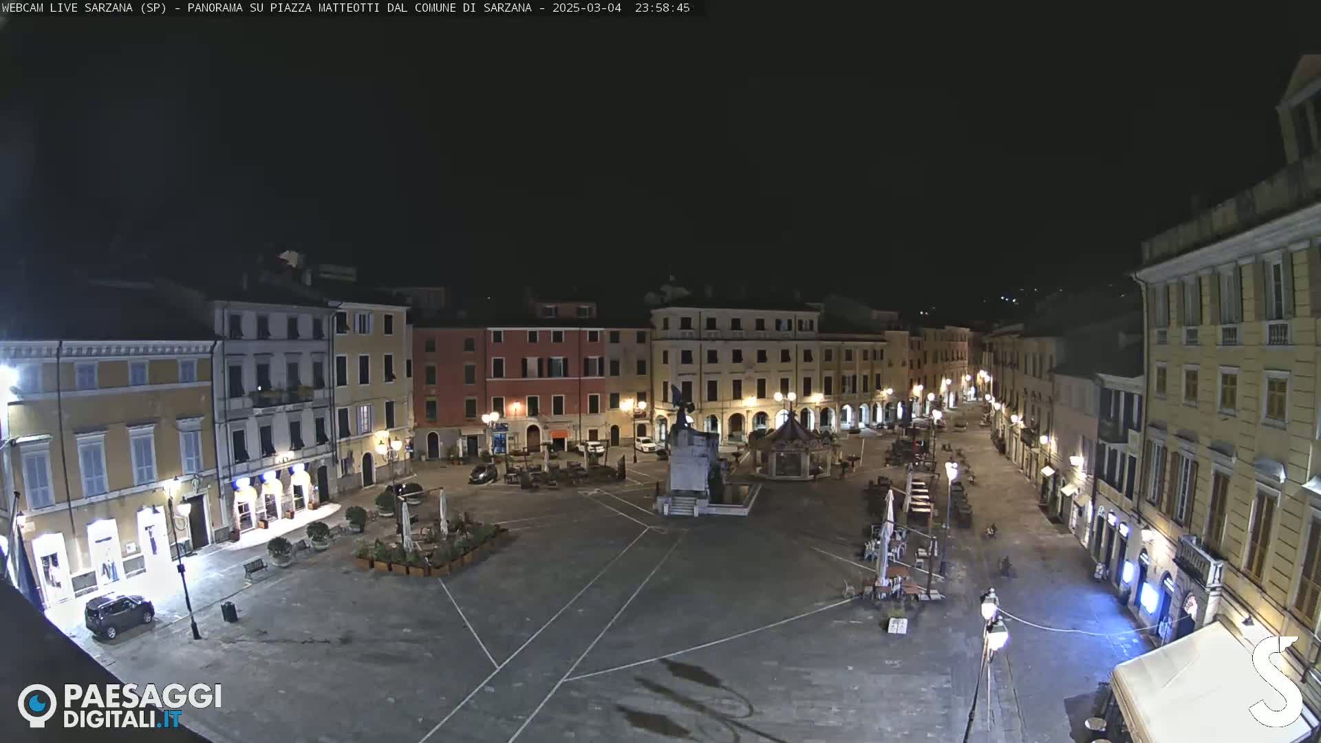 A nighttime, wide shot of a town square with buildings surrounding it, illuminated by streetlights.