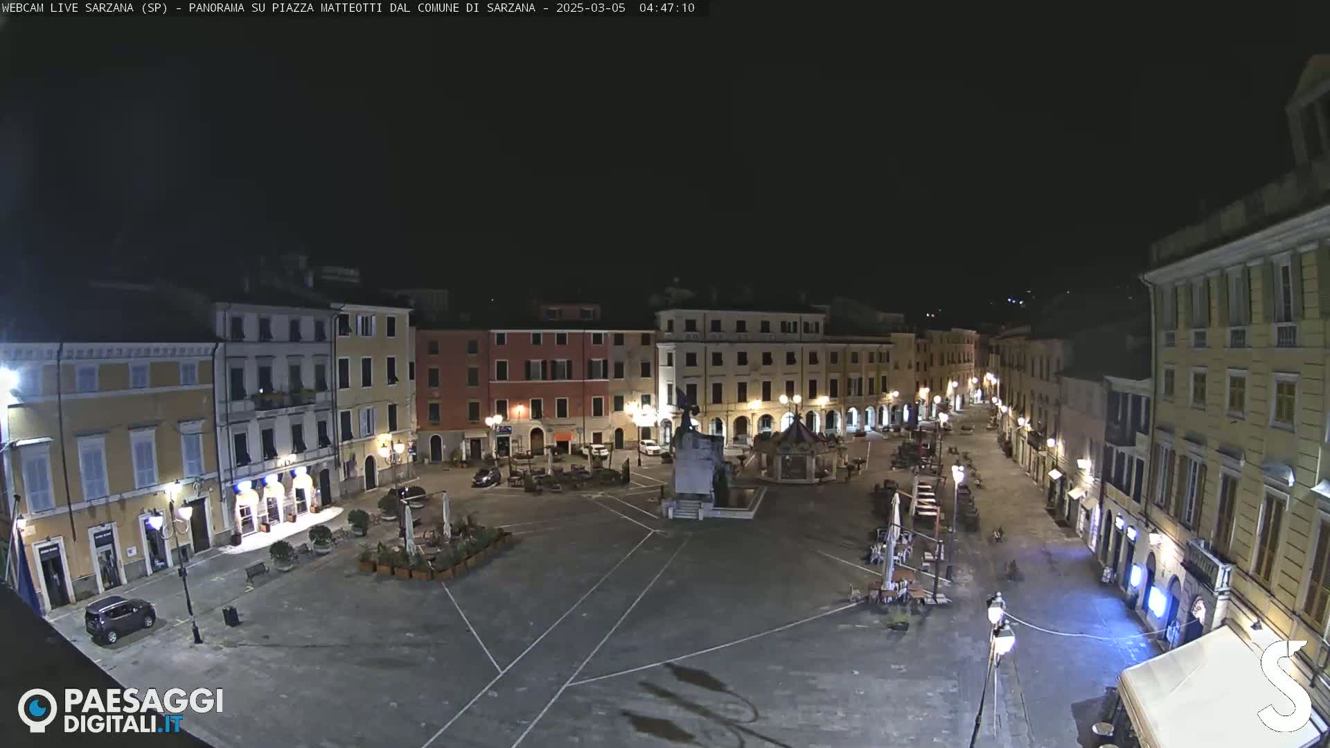 A mostly empty town square at night, surrounded by buildings with lights illuminating the area.
