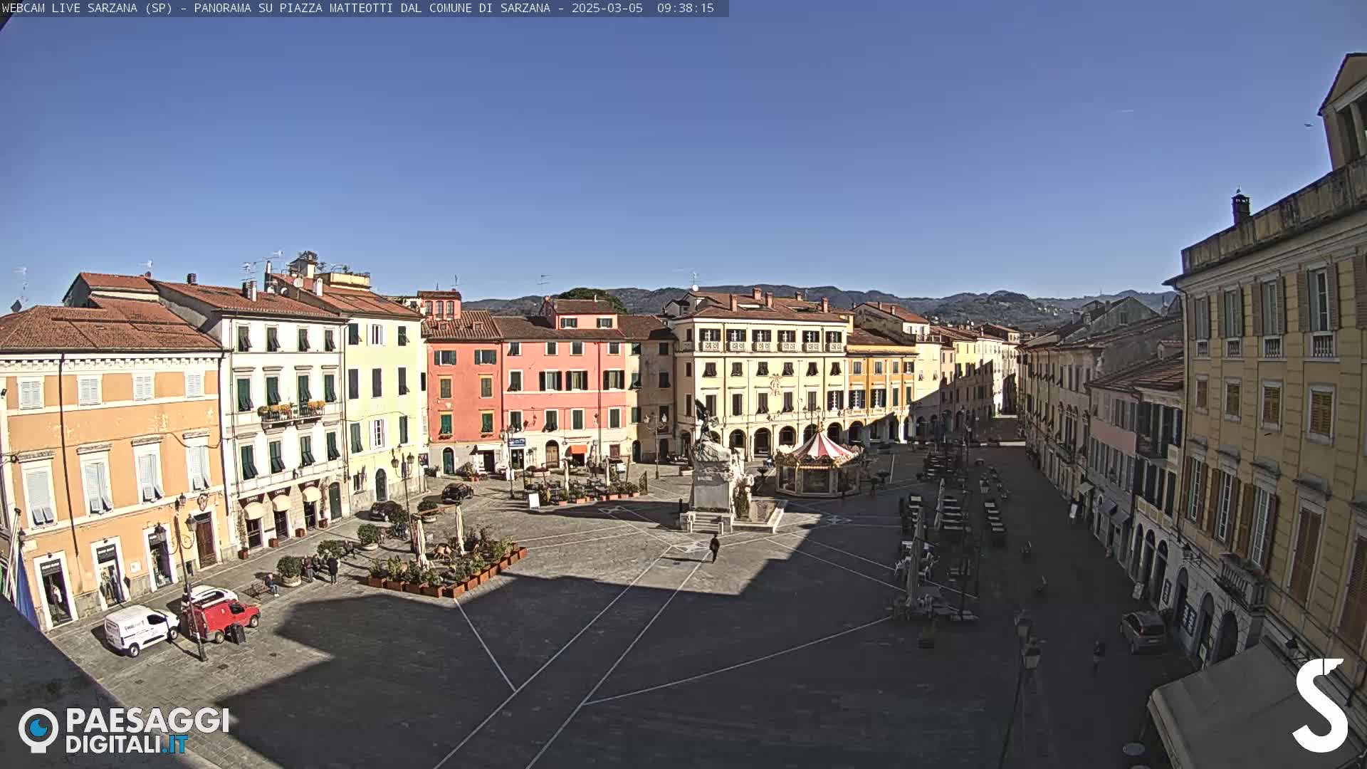 A sunny day reveals a large, open town square surrounded by multi-story buildings, featuring a monument and a carousel in the center.