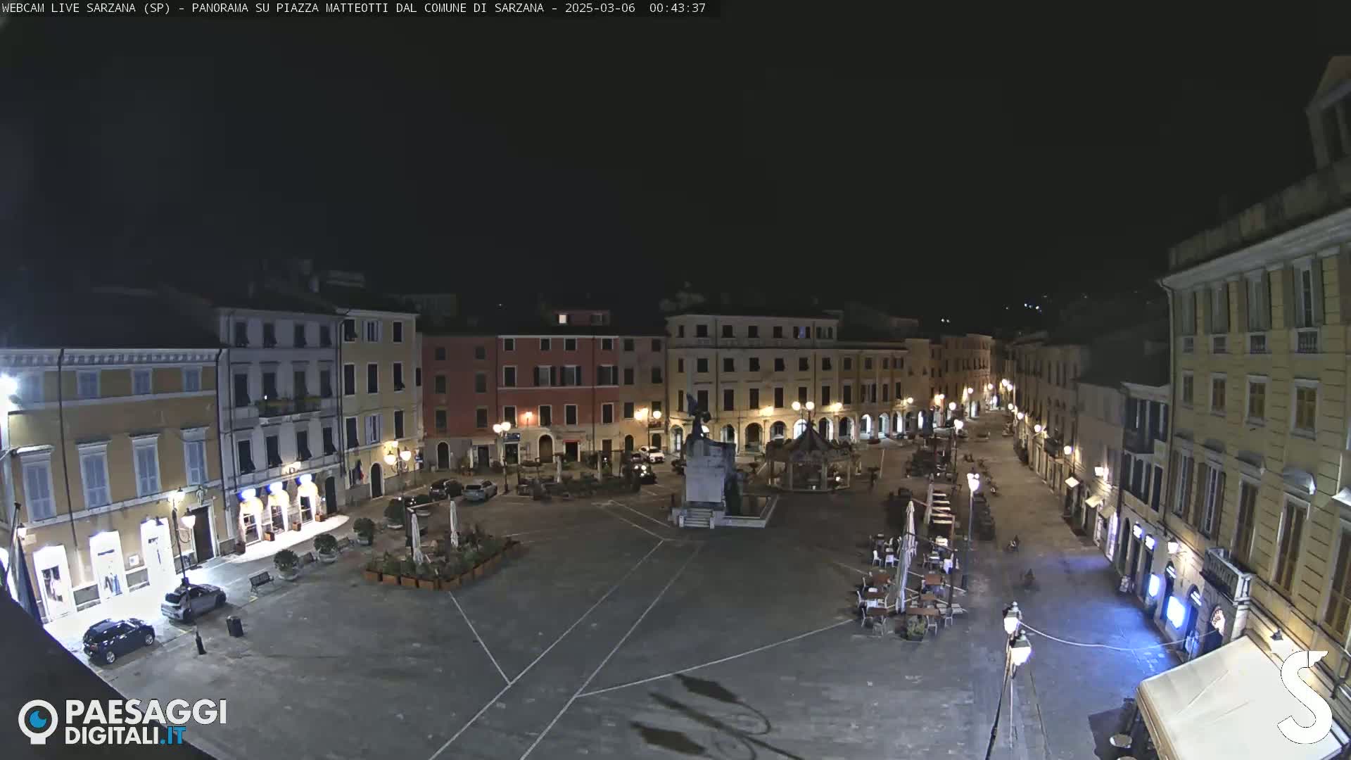 A nighttime, high-angle view of a town square with several buildings surrounding it, a few cars parked, and some outdoor seating.