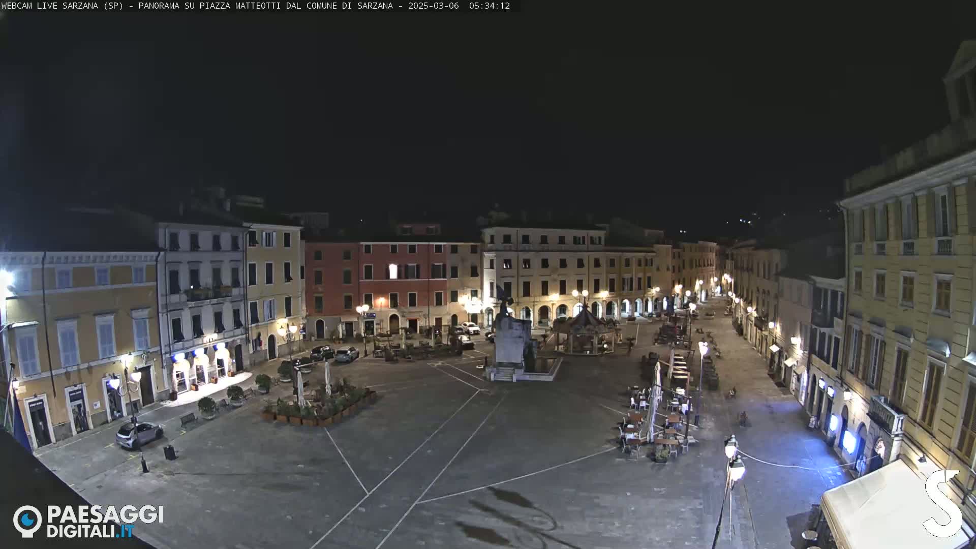 A nighttime, high-angle view of a mostly empty town square surrounded by buildings with illuminated windows and streetlights.