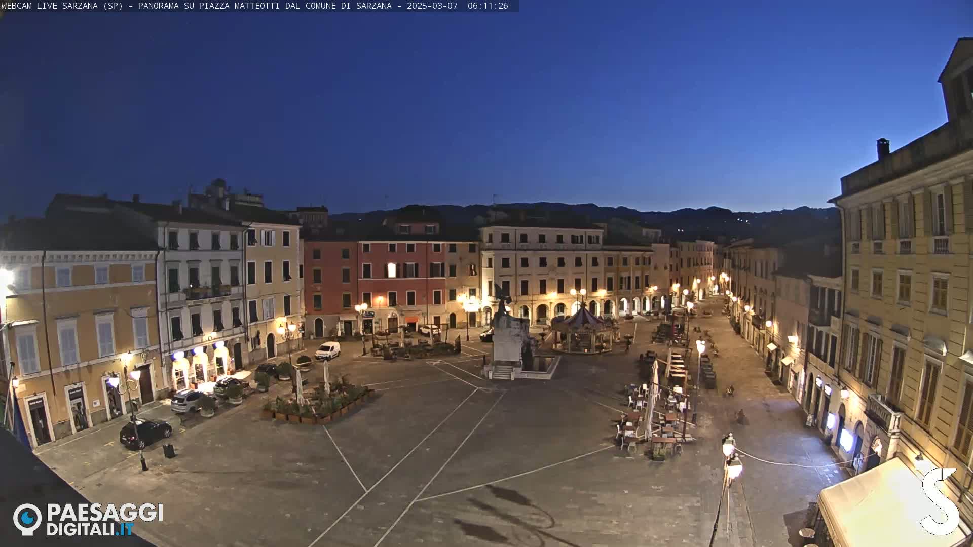 A mostly empty town square at twilight, surrounded by illuminated buildings, under a clear dark blue sky.