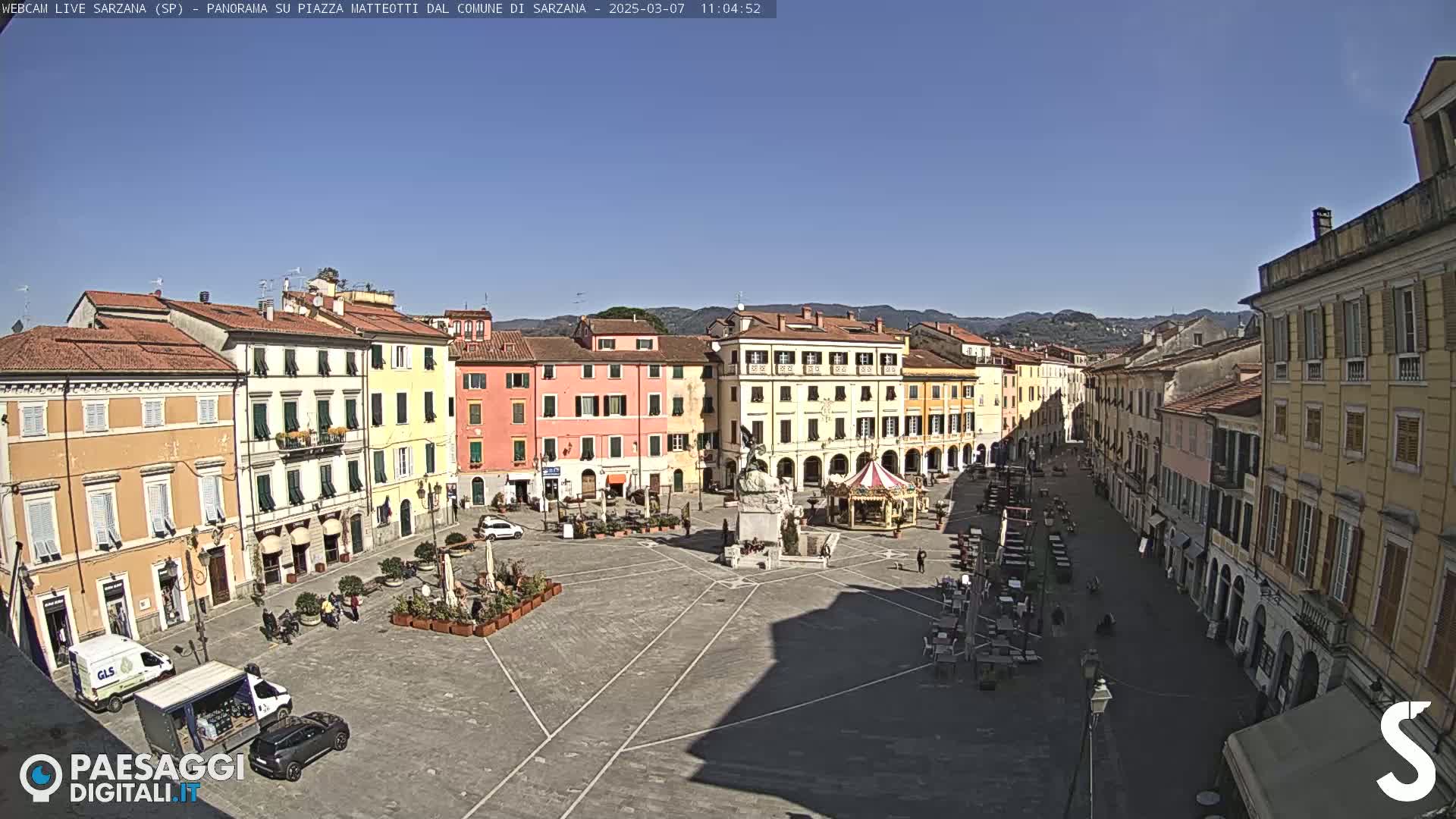 A large town square, surrounded by multi-story buildings under a clear, sunny sky, contains a few people, vehicles, and outdoor seating areas.