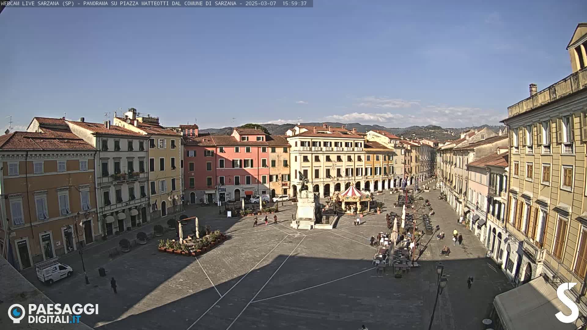 A sunny day reveals a large town square surrounded by multi-story buildings, with a few people and a carousel visible.