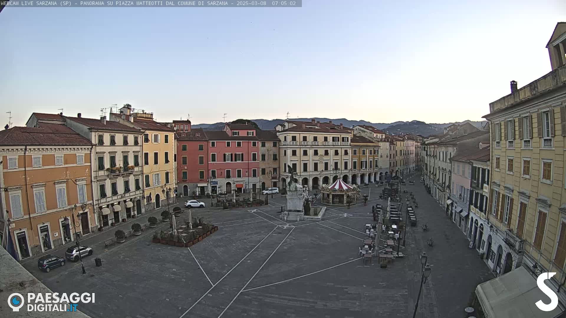 A mostly empty town square with several multi-story buildings surrounding it under a clear, light-blue sky.