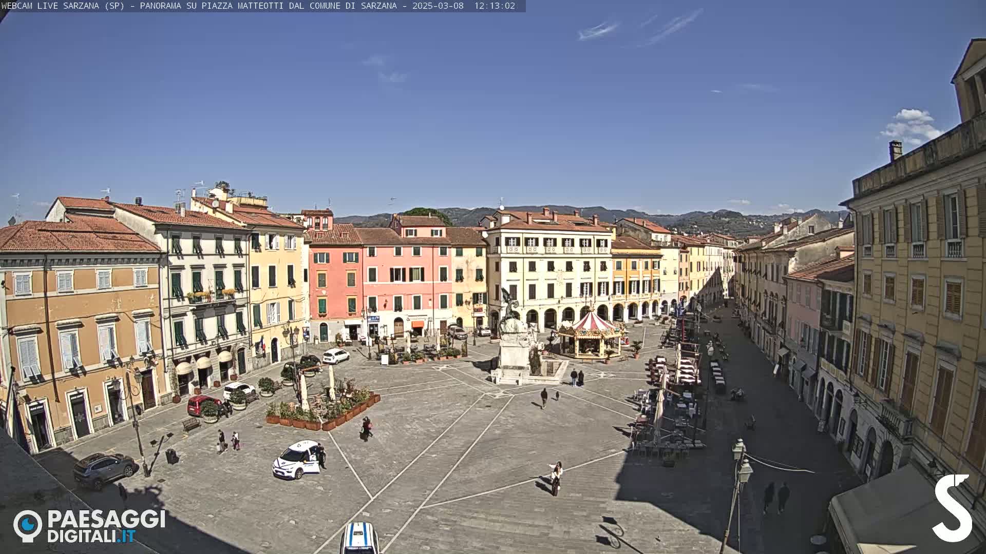 A sunny day reveals a large town square surrounded by multi-story buildings, a monument, a carousel, and a few people and vehicles.
