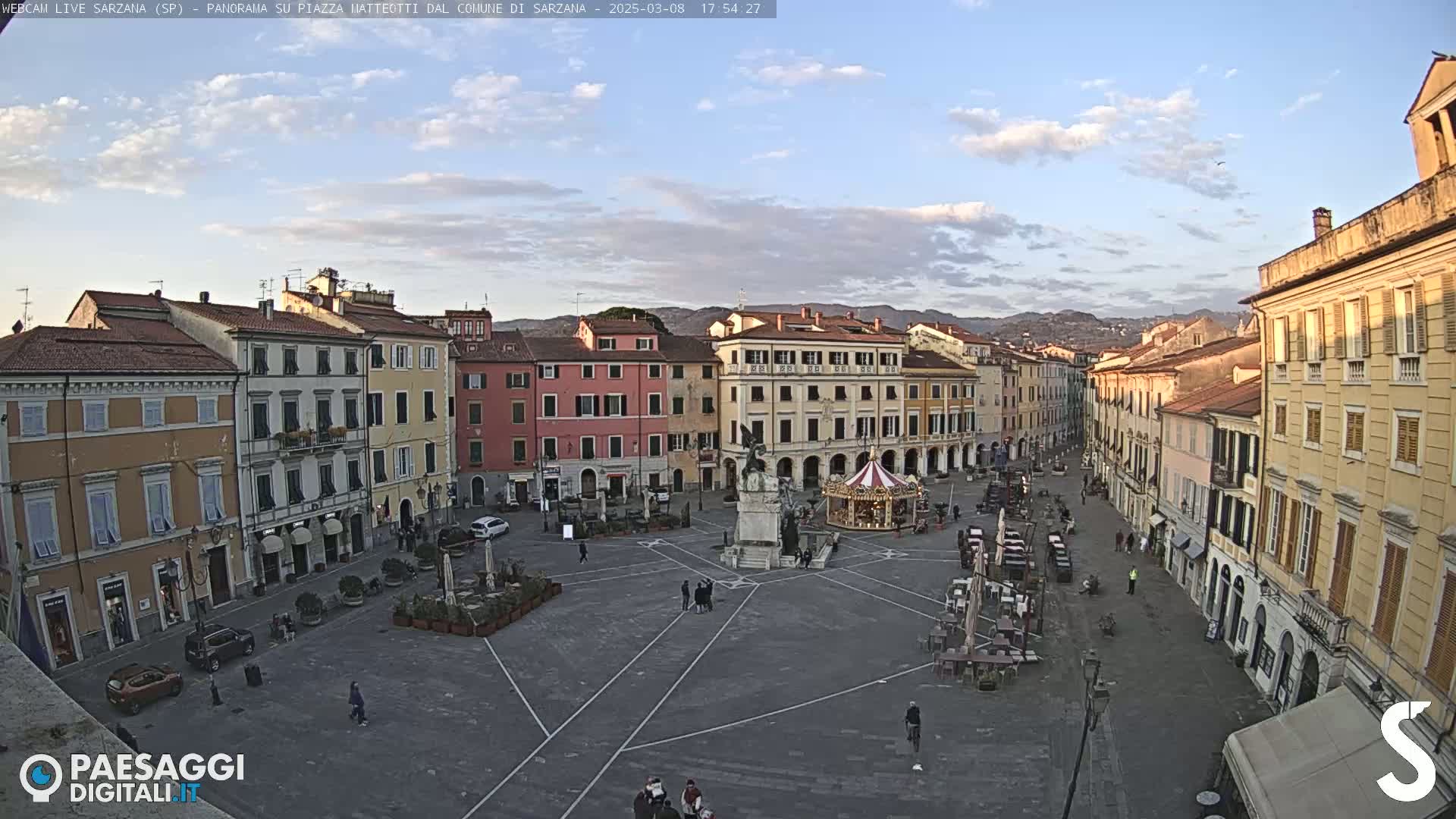 A mostly empty town square, surrounded by multi-story buildings, under a partly cloudy sky.