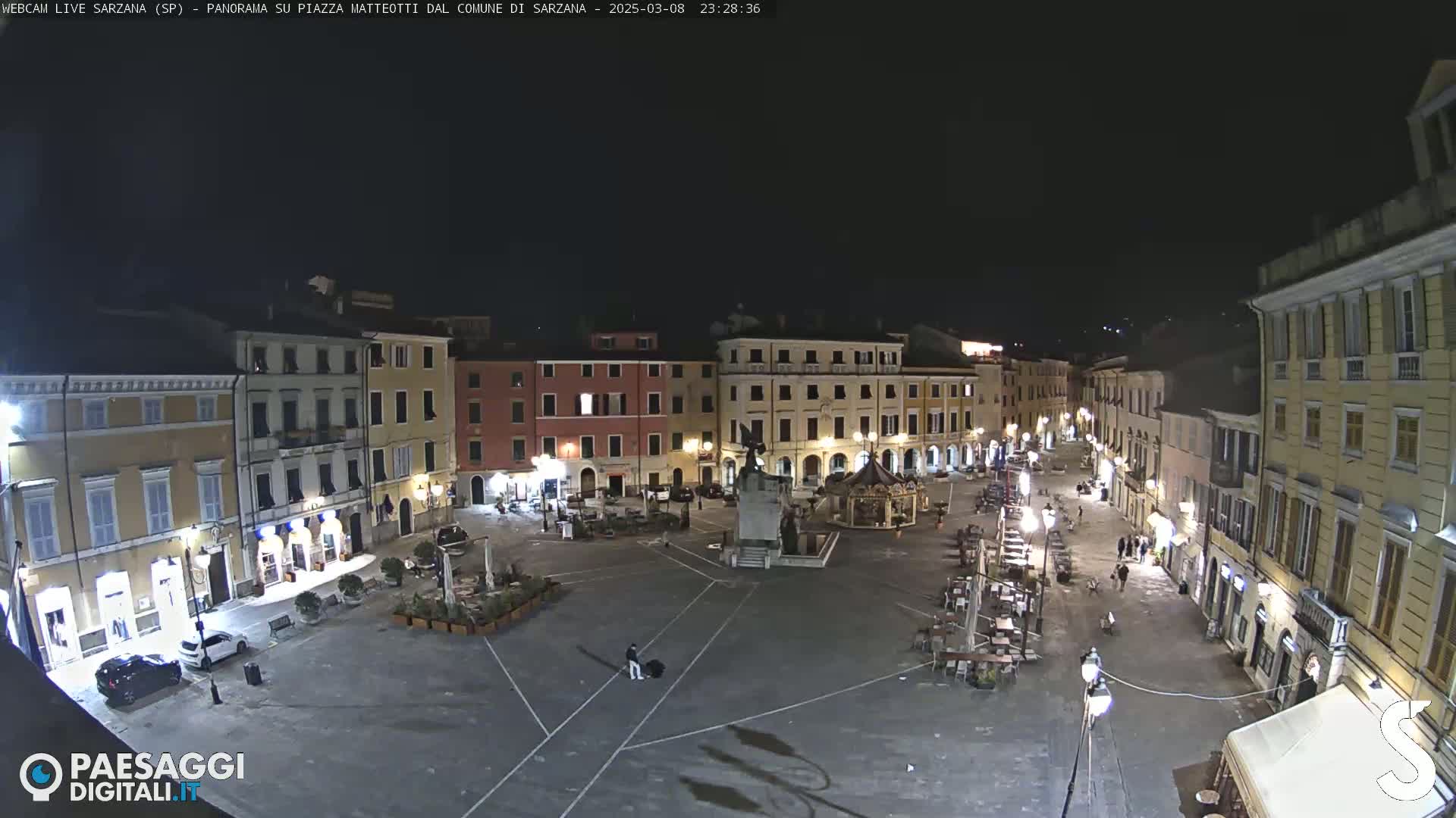 A nighttime, high-angle view of a sparsely populated town square surrounded by multi-story buildings, with outdoor seating at several cafes and restaurants.