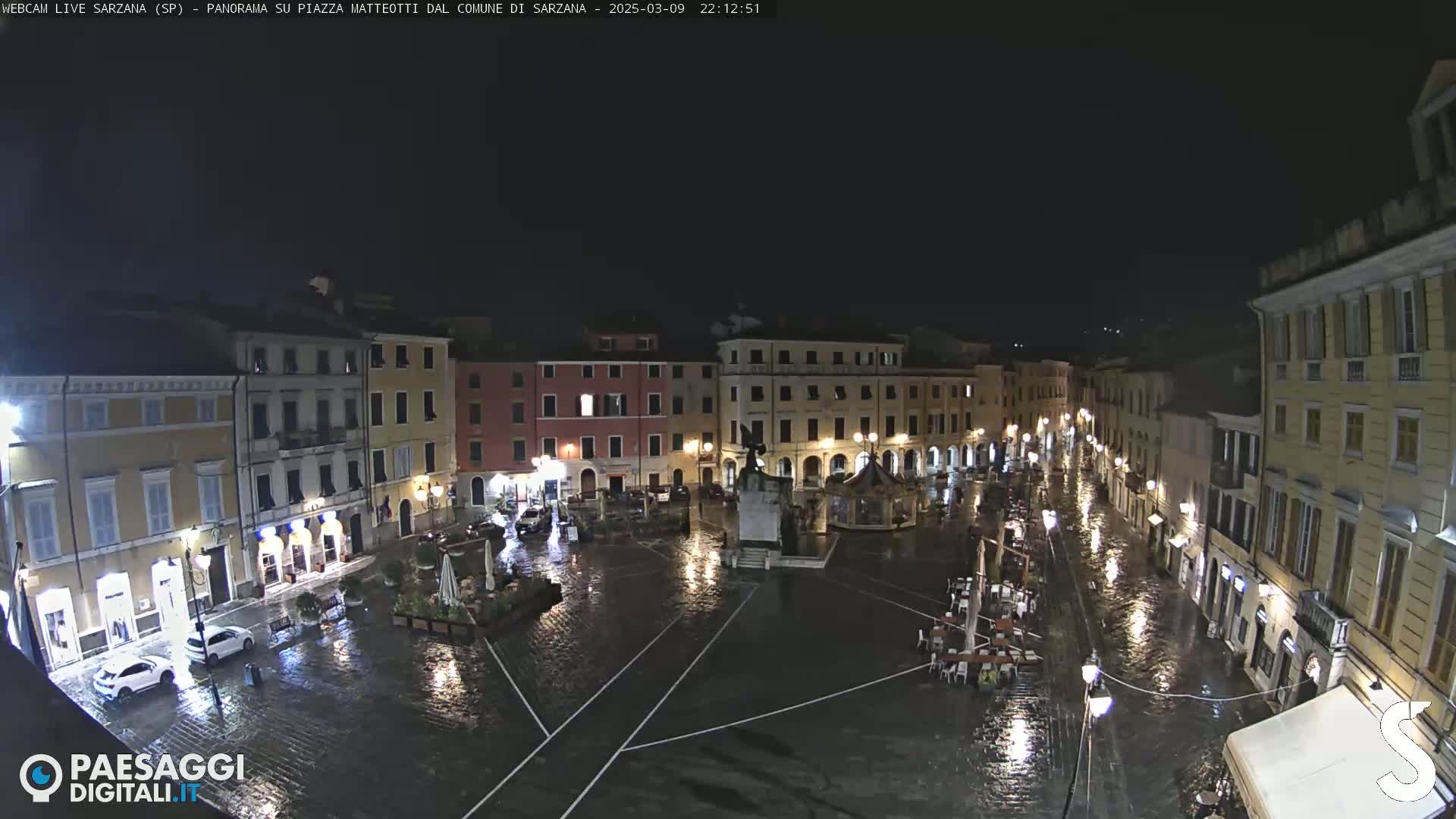 A rain-soaked town square at night, surrounded by illuminated buildings.