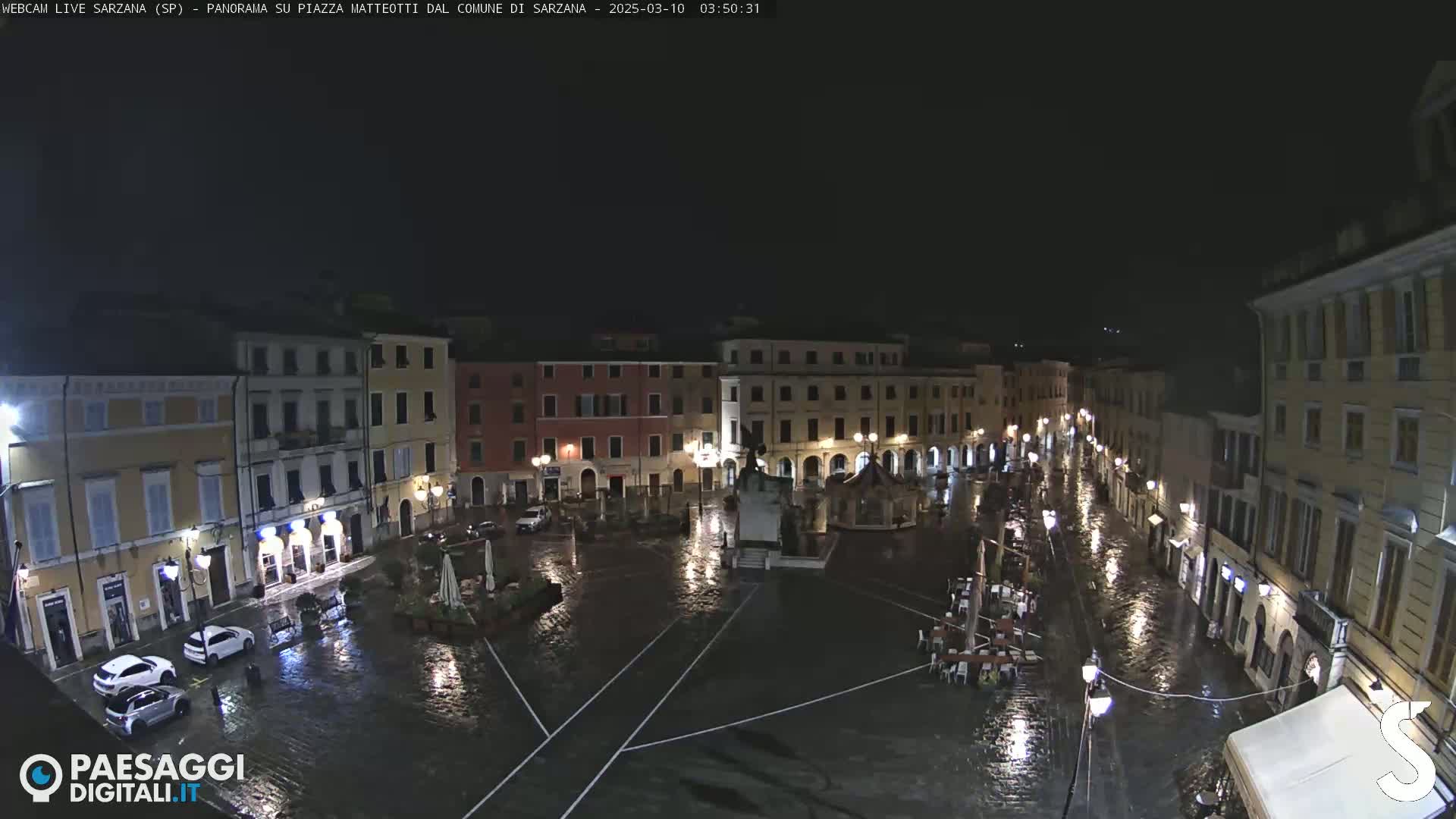 A rain-soaked town square at night, surrounded by buildings with illuminated windows, is viewed from above.