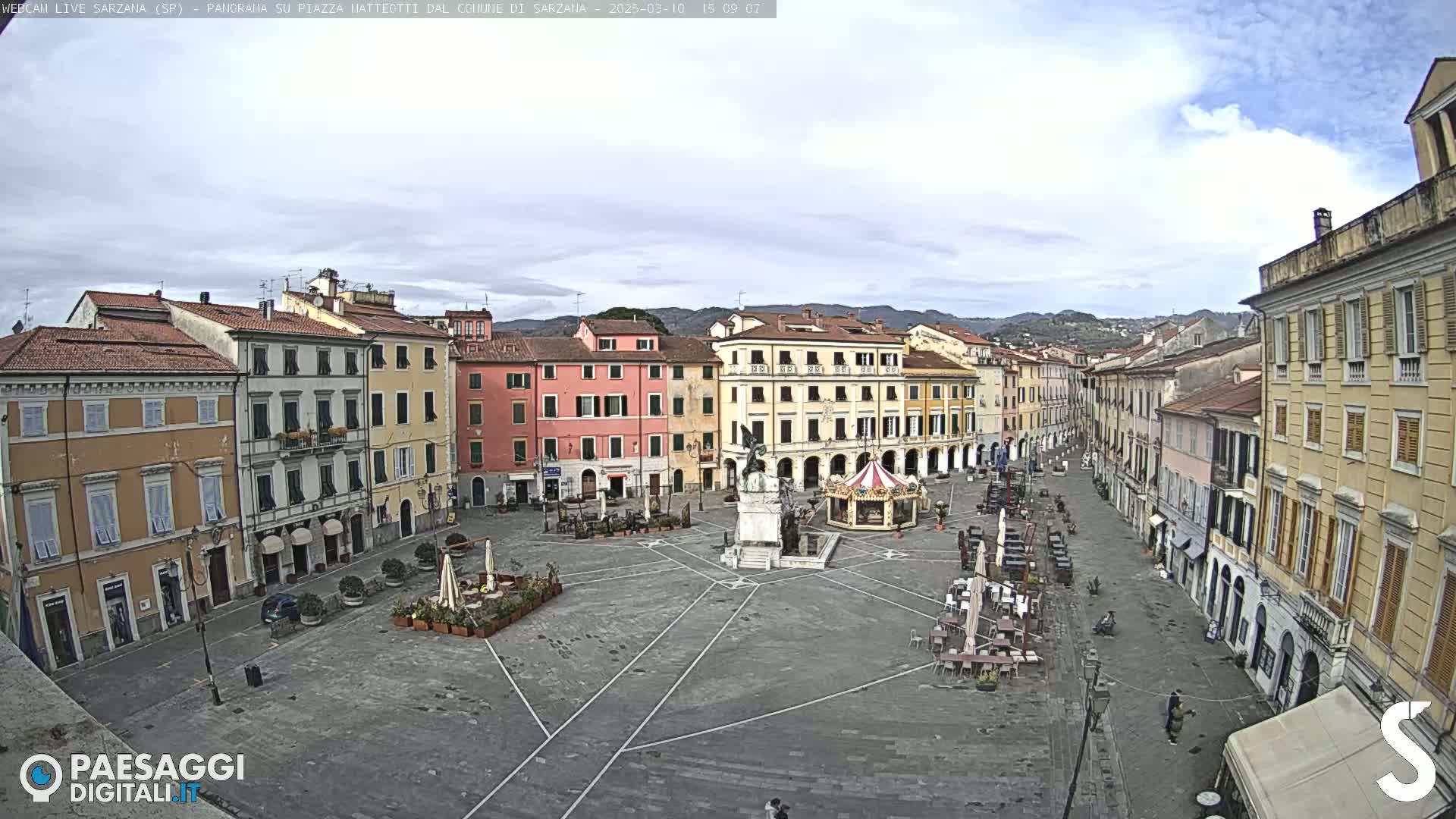A mostly empty town square with several multi-story buildings surrounding it under a partly cloudy sky.