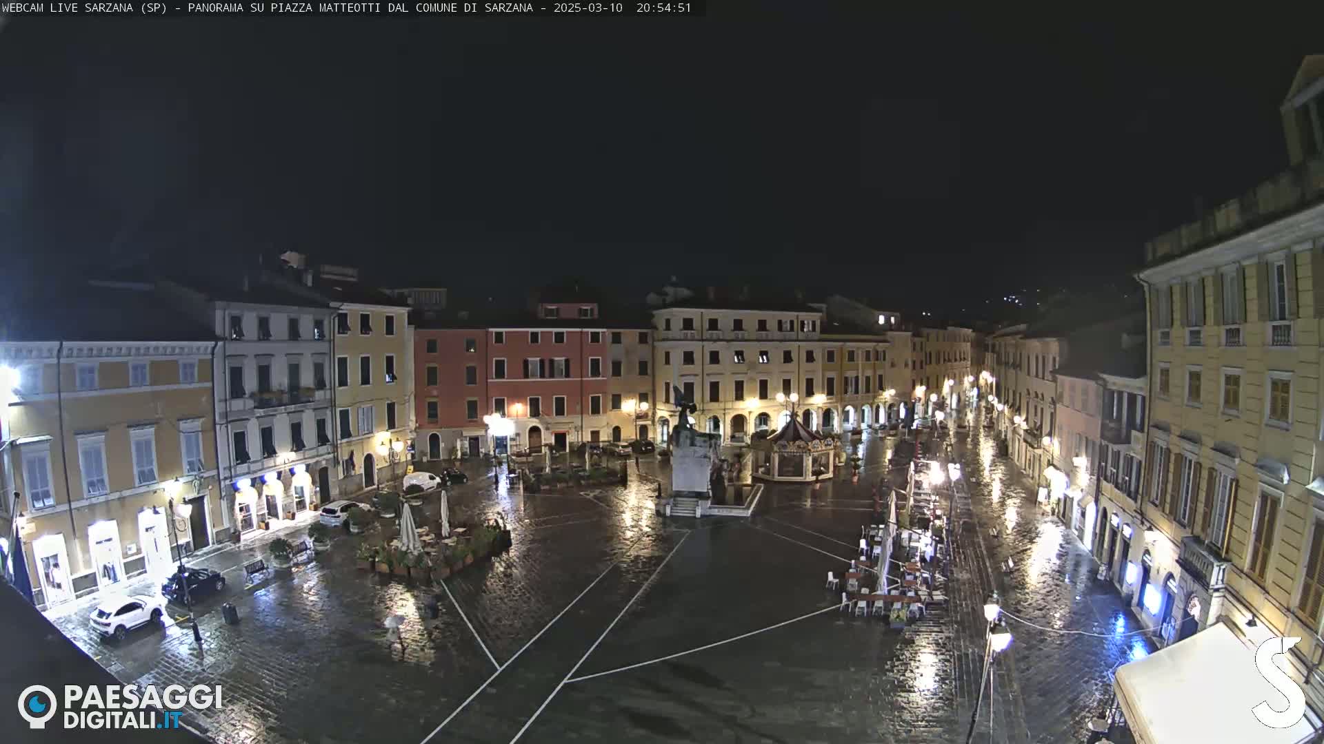 A rain-slicked town square at night, illuminated by streetlights, is surrounded by buildings.