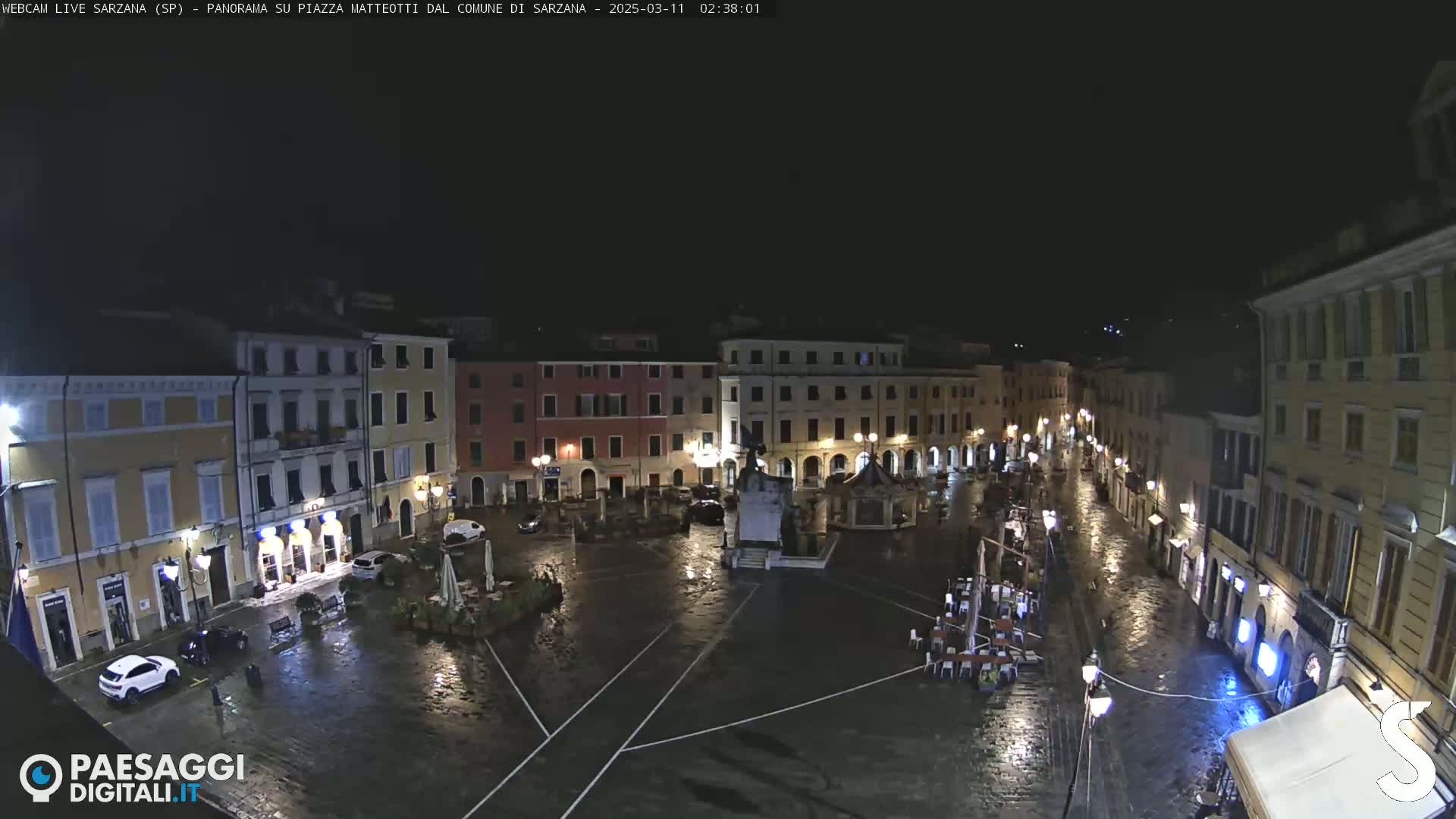 A rain-slicked, nighttime town square is illuminated by streetlights, showing several buildings surrounding a central monument and a few parked cars.