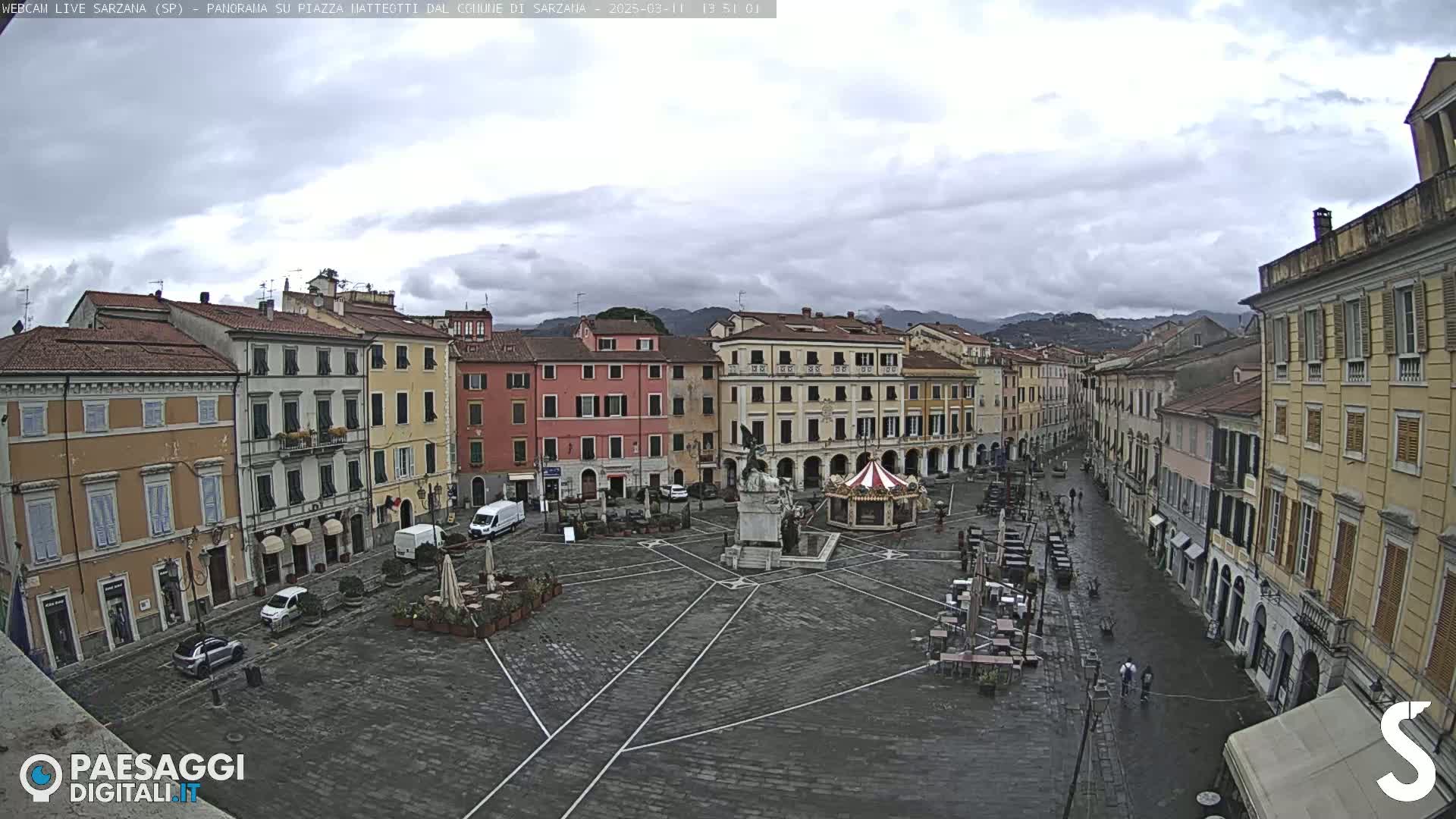 A mostly empty town square with surrounding buildings under an overcast sky is shown.