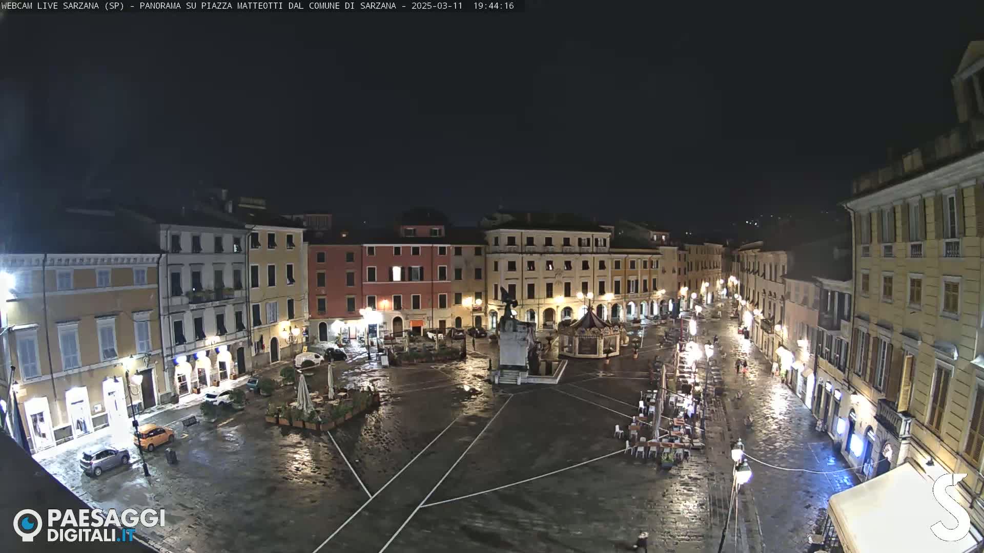 A wet, nighttime view of a town square surrounded by illuminated buildings.