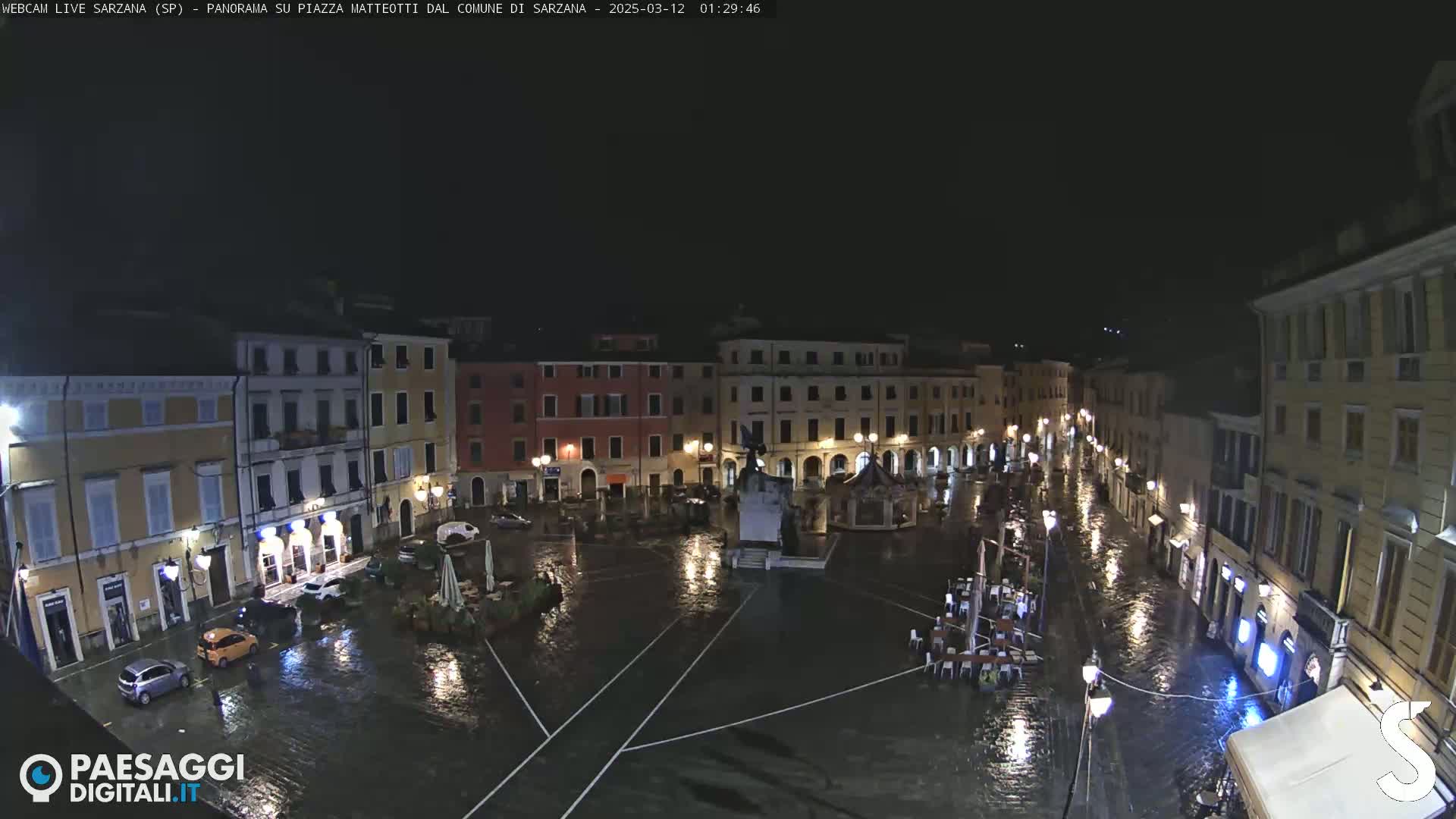 A rain-soaked town square at night, surrounded by buildings with illuminated windows, features a central monument and several cars parked in the flooded plaza.