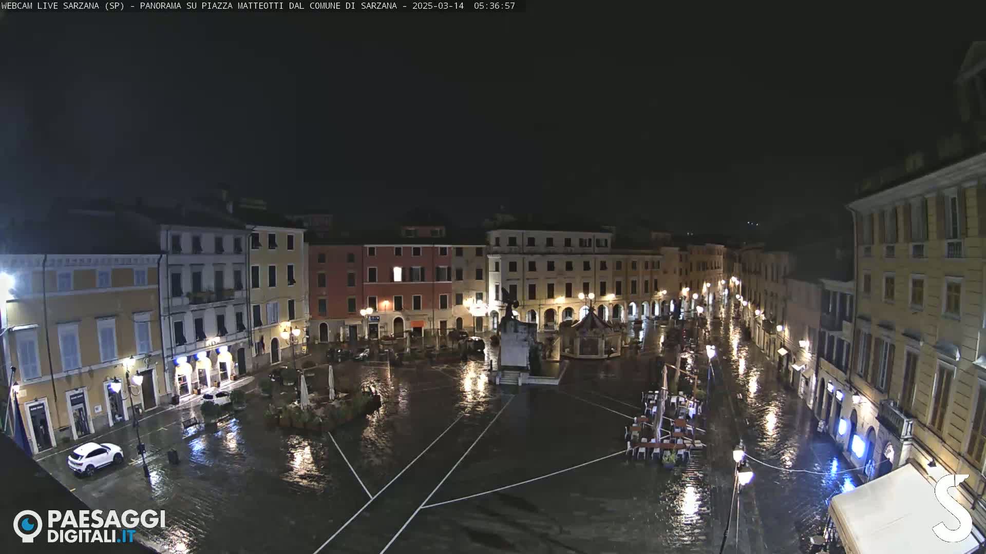 A nighttime, rain-flooded town square is surrounded by buildings with illuminated windows and streetlights, reflecting in the standing water.