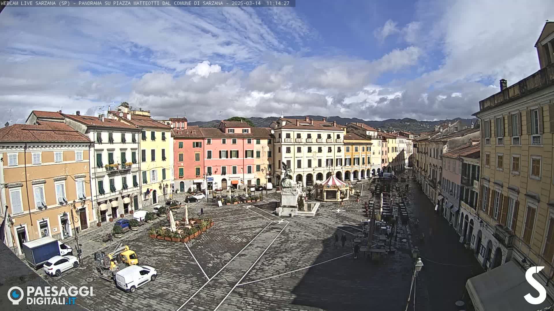 A mostly sunny day reveals a large, cobblestone town square surrounded by multi-story buildings, with a few cars and some outdoor seating visible.