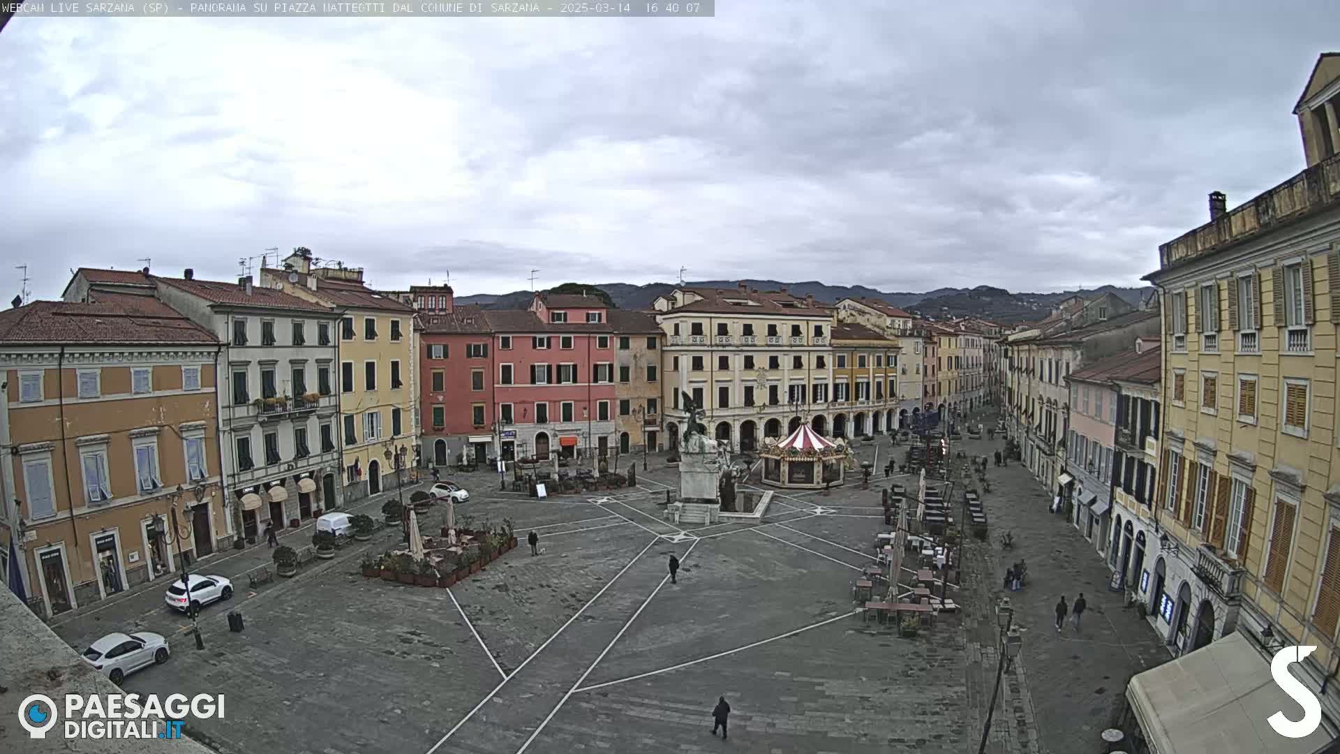 A mostly empty town square, surrounded by multi-story buildings, under an overcast sky.