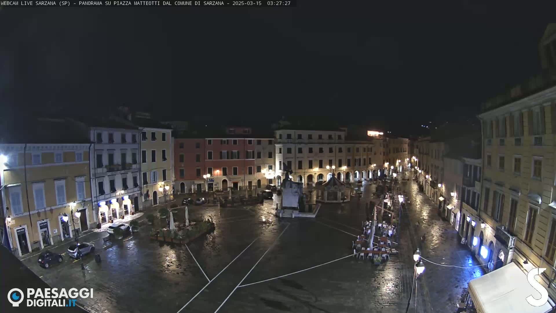 A rain-slicked, nighttime town square is sparsely populated with a few cars and some outdoor furniture; buildings surround the square, their lights illuminating the wet cobblestones.