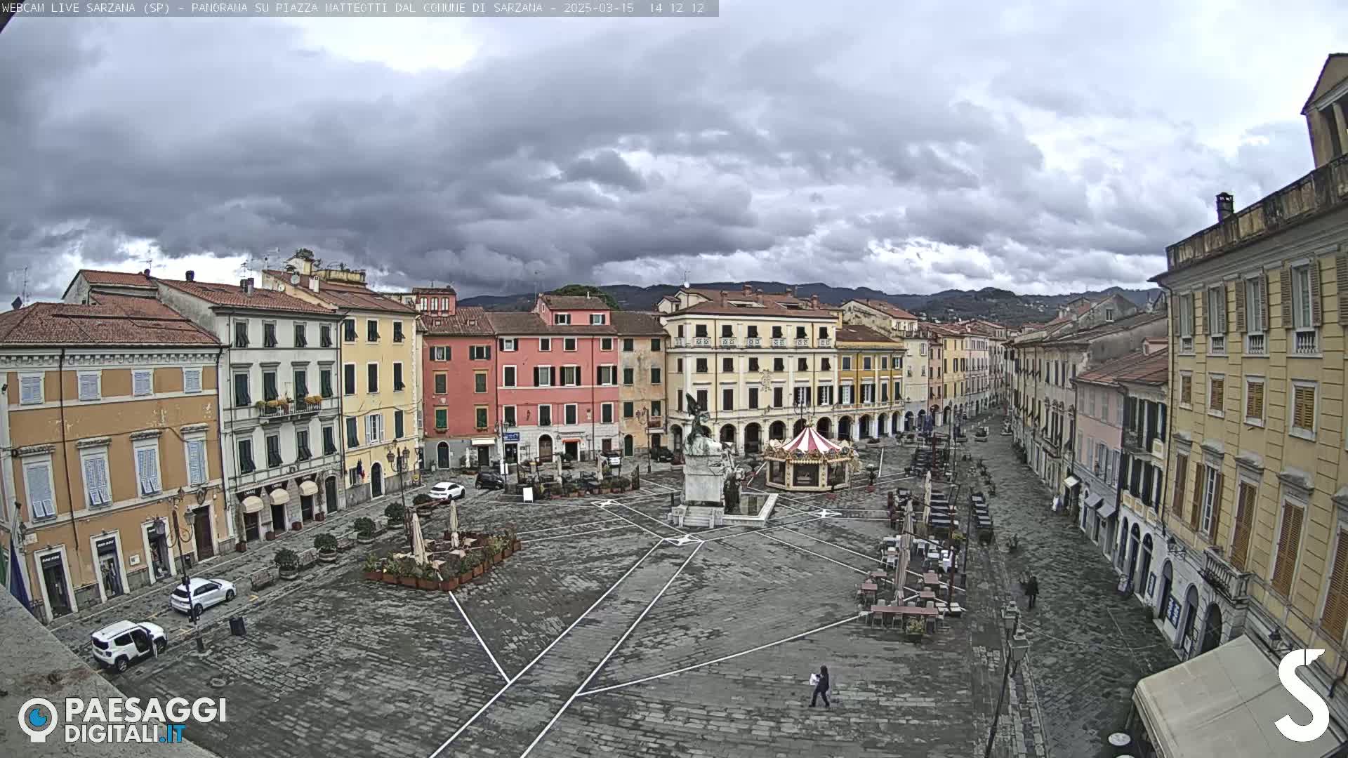 A mostly empty town square with several buildings surrounding it under a cloudy sky.