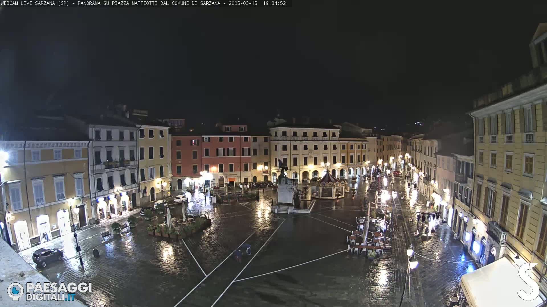 A rain-soaked town square at night, illuminated by streetlights, is surrounded by multi-story buildings.