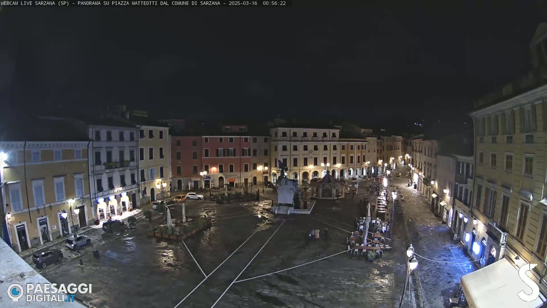 A wet, nighttime view of a town square with several buildings surrounding it, sparsely populated with people and cars.