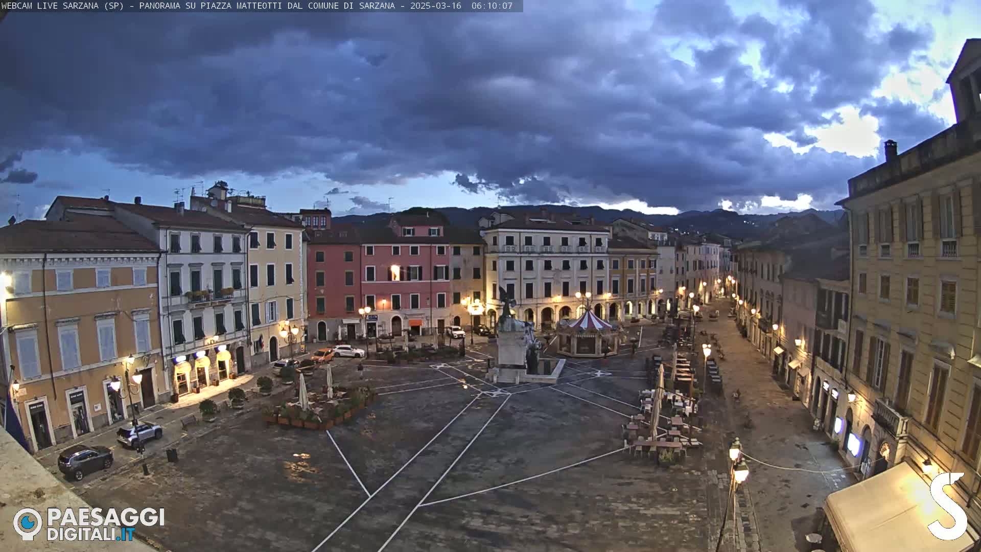 A mostly empty town square at twilight under a cloudy sky, surrounded by buildings with outdoor seating areas.