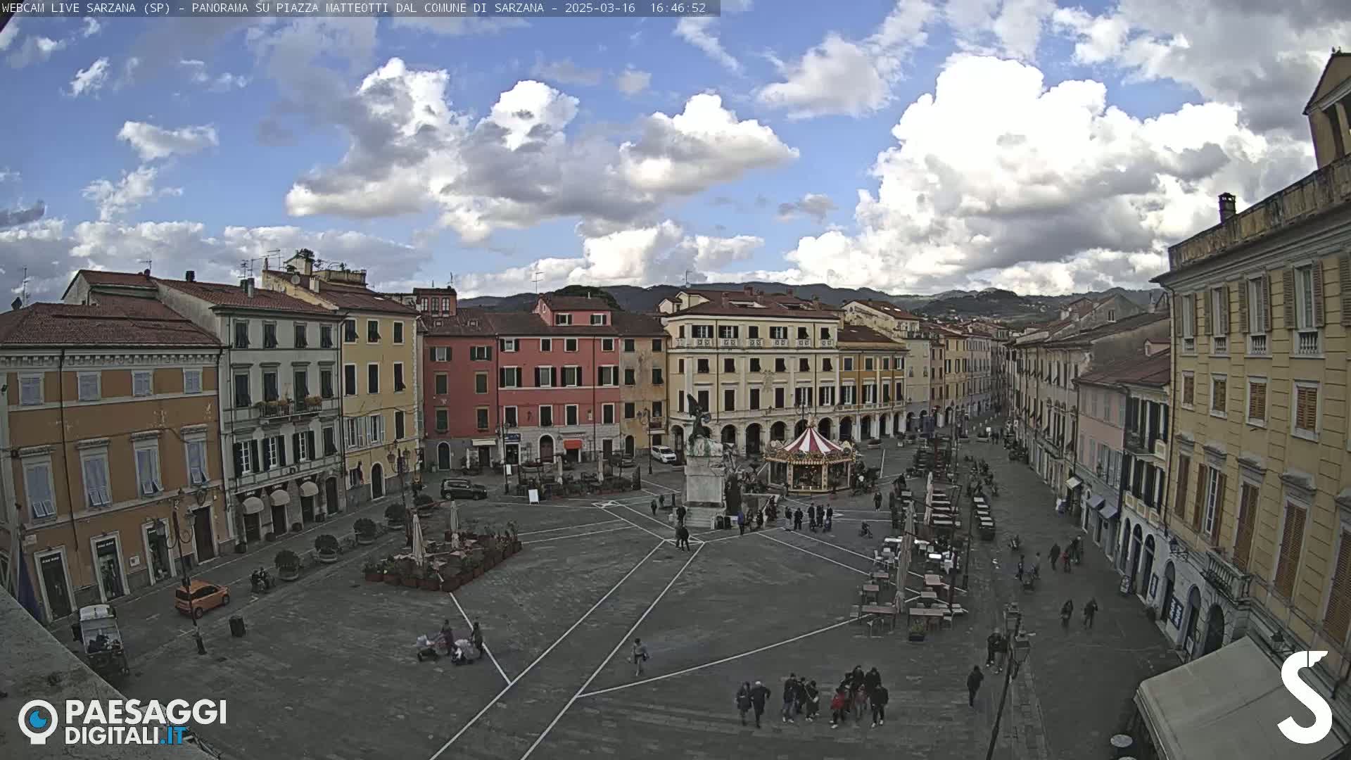 A partly cloudy day overlooks a large town square with several buildings, a carousel, and people walking around.
