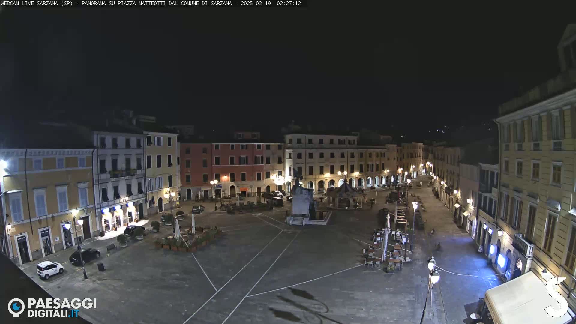 A nighttime, aerial view of a mostly empty town square, surrounded by illuminated buildings.