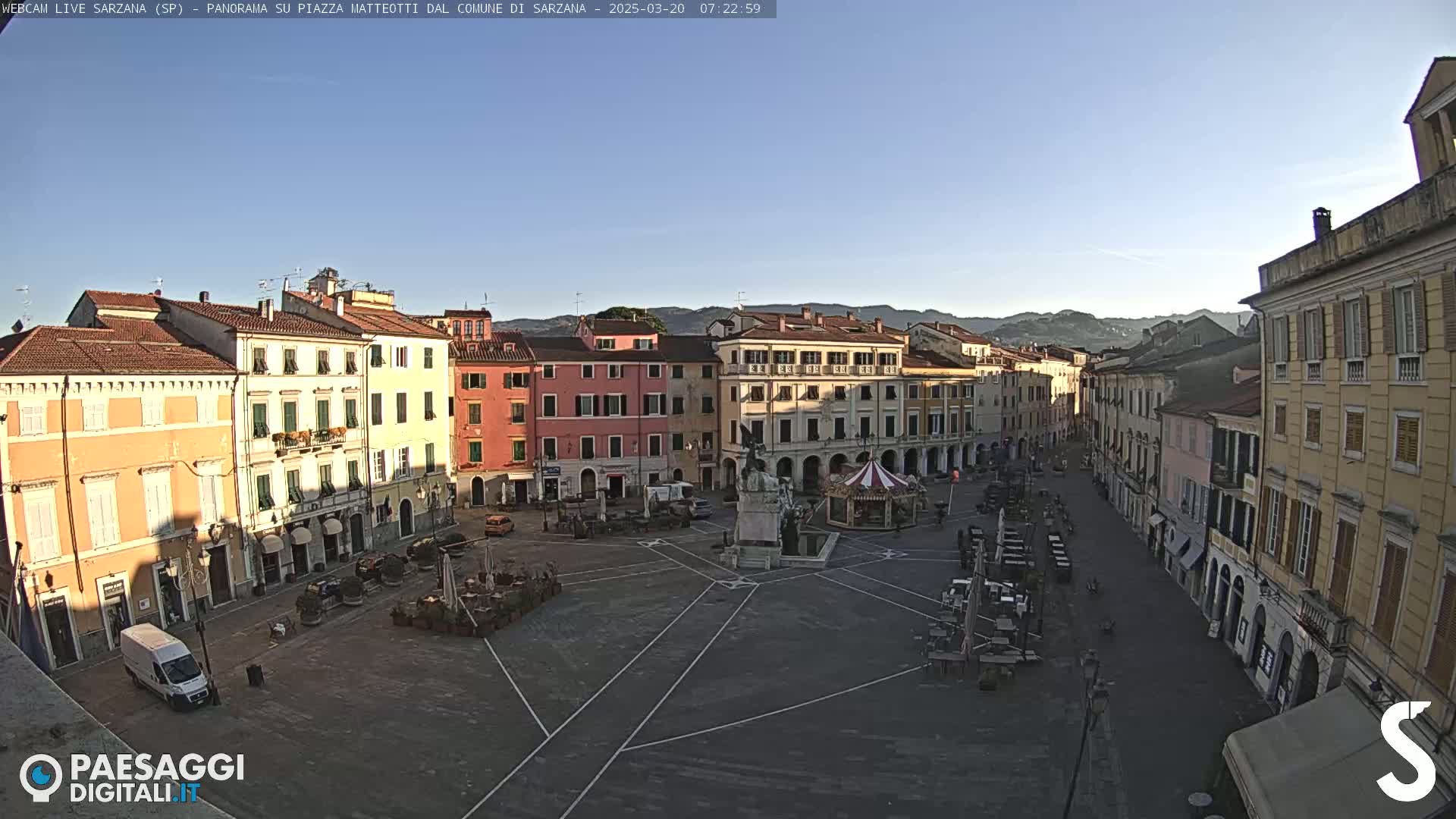 A mostly empty town square, surrounded by multi-story buildings, under a clear, bright morning sky.