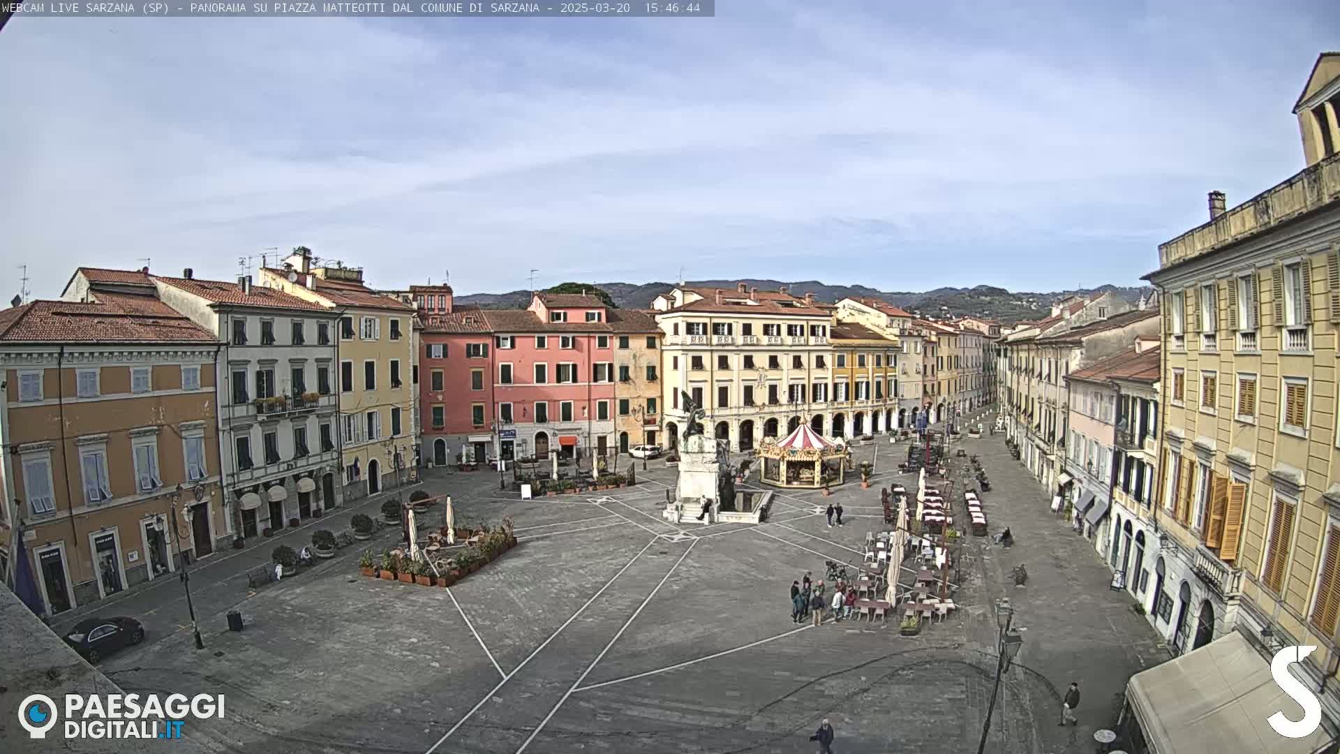 A mostly sunny day overlooks a large town square in Italy, featuring several multi-story buildings, a monument, and a carousel.