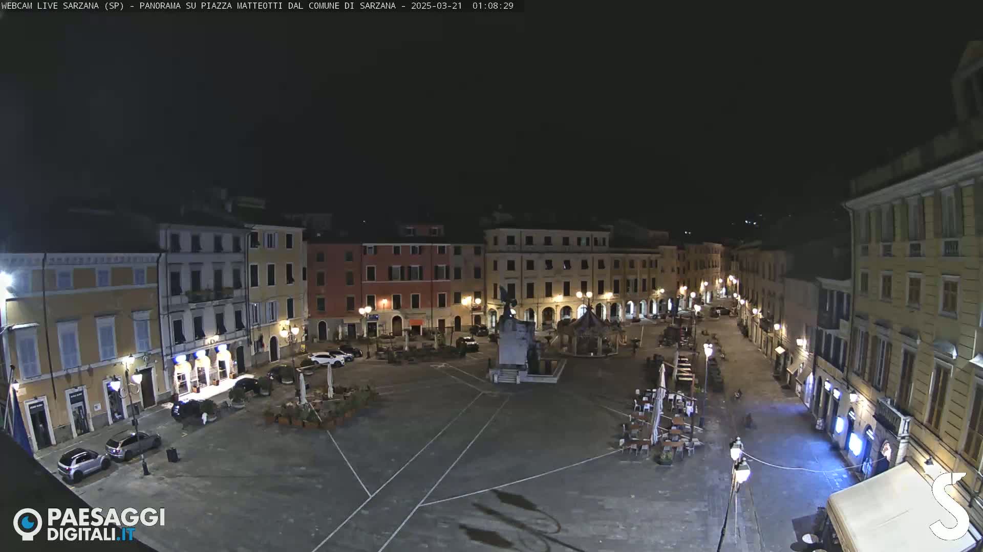 A nighttime, high-angle view of a mostly empty town square with several buildings surrounding it, featuring some cars parked and outdoor seating areas with tables and chairs.