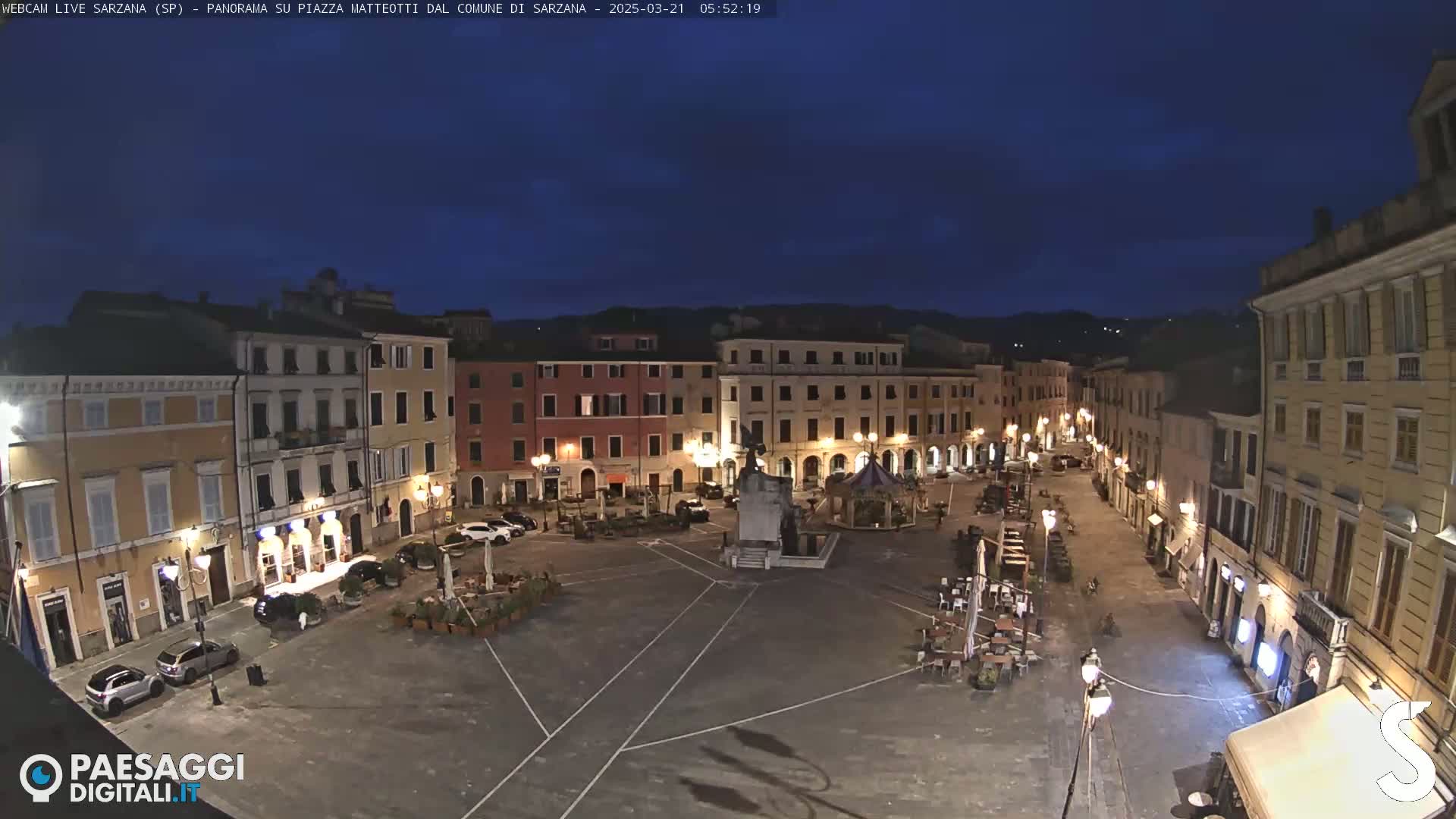 A mostly empty town square at night, illuminated by streetlights, is surrounded by buildings under a dark blue, twilight sky.