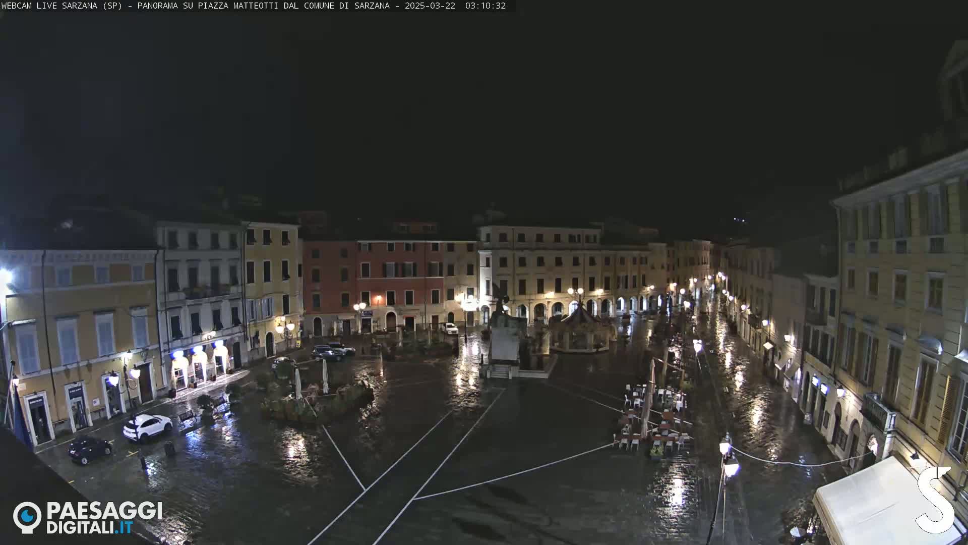 A rain-soaked town square at night, surrounded by illuminated buildings, with a few cars and outdoor seating visible.